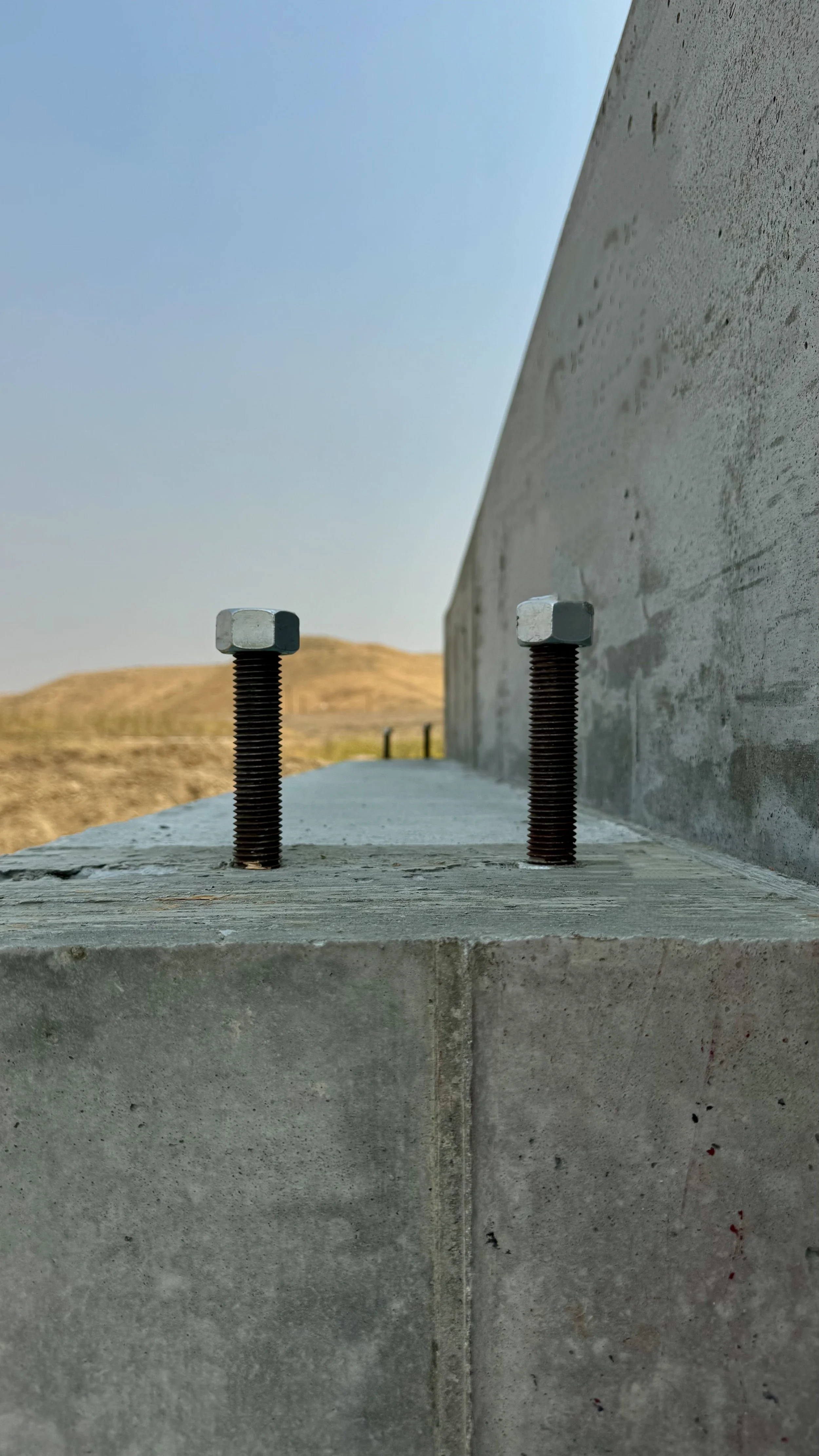 Close-up of two metal bolts protruding from a concrete surface with a cloudy sky and barren landscape in the background.