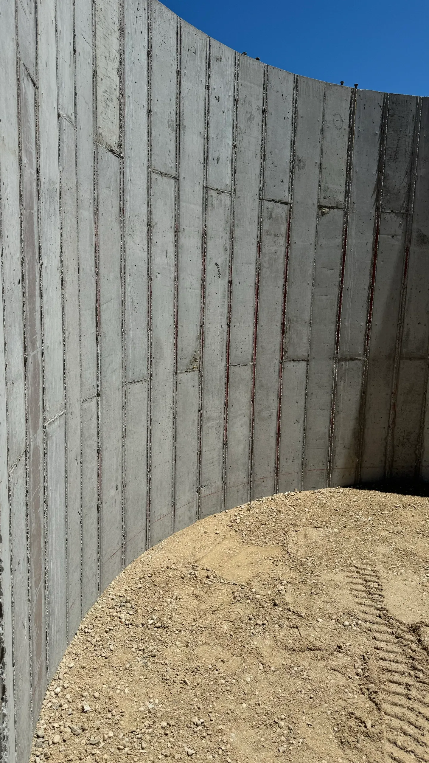 A curved concrete segment wall under construction with dirt ground in front and a clear blue sky above.
