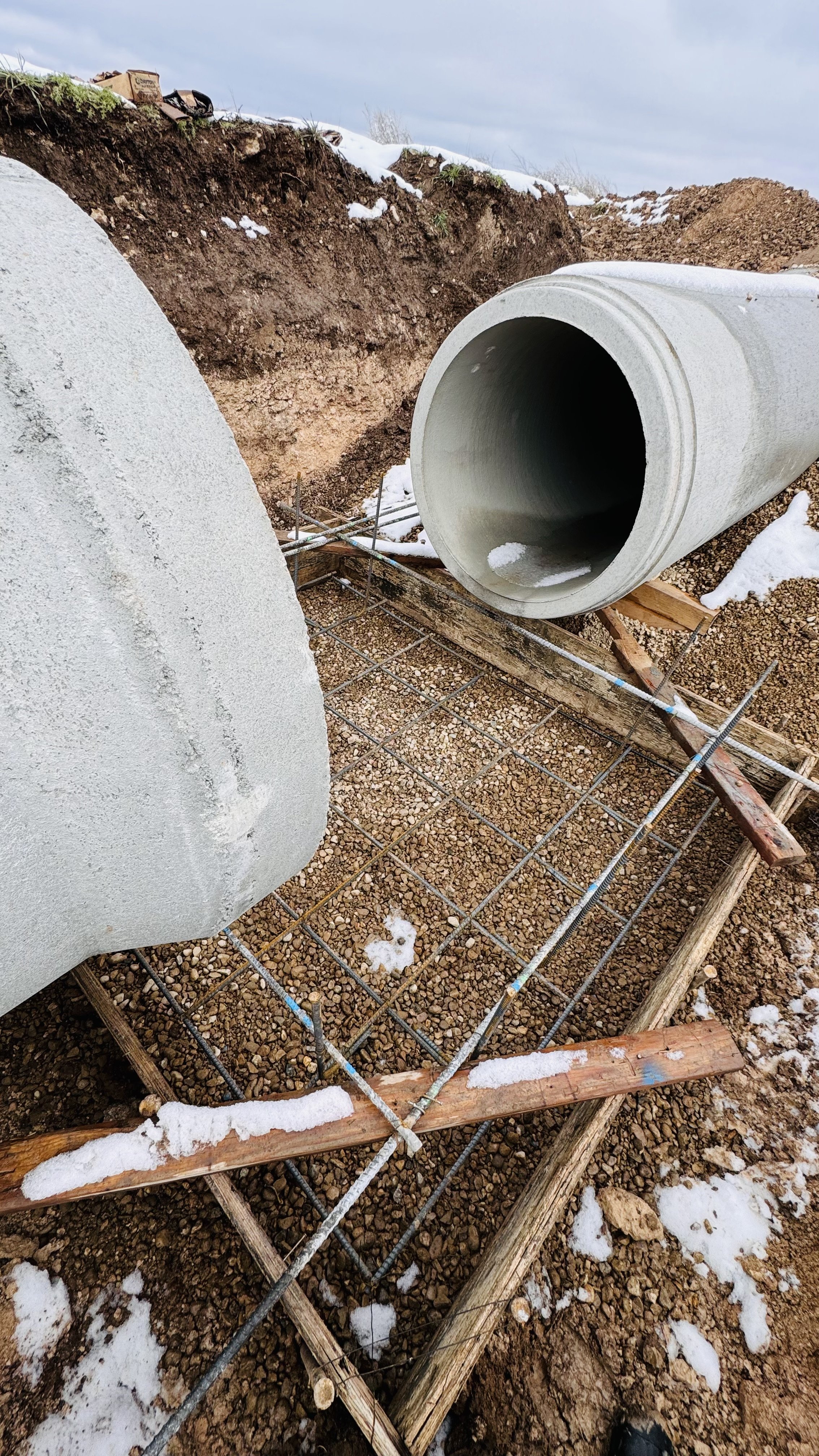 Construction site with large concrete pipe, rebar, wooden formwork, and exposed dirt and snow.