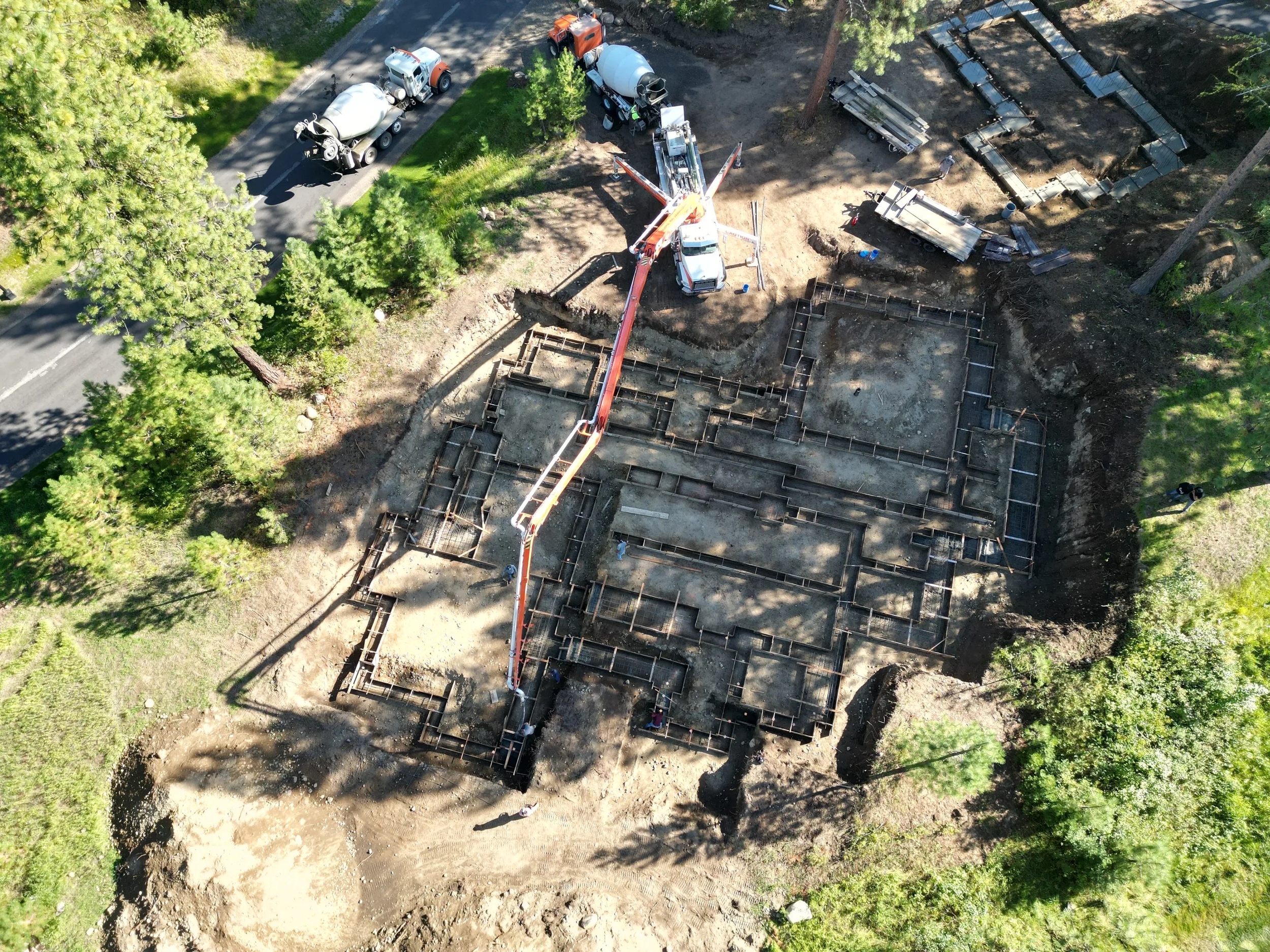 An aerial view of a construction site where the foundation is being prepared. There are concrete mixer trucks on the road nearby, and construction equipment like a crane and scaffolding around the excavation area. Trees surround the site.