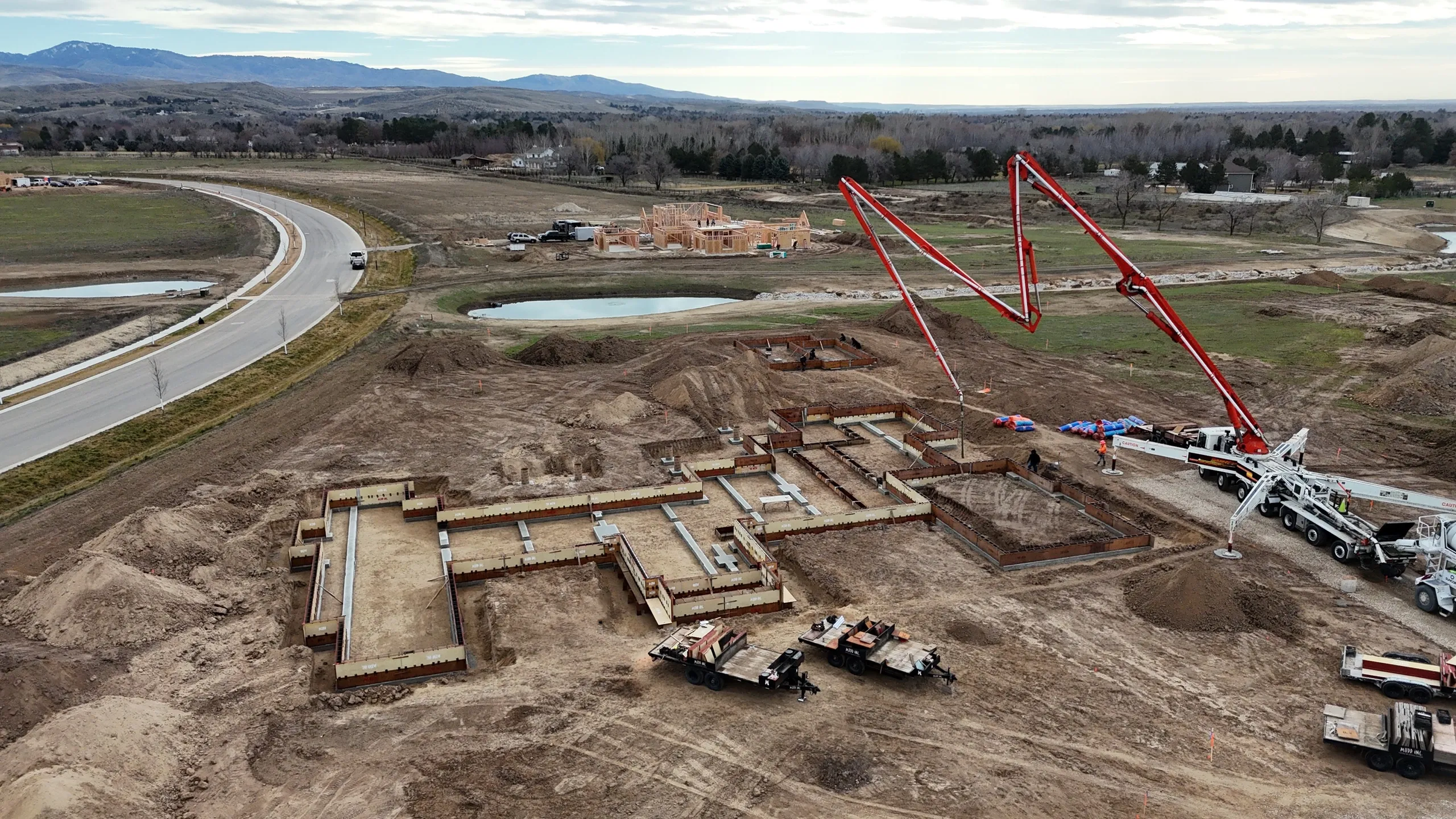 Construction site with foundation and concrete pouring, two large red concrete pump trucks, and a partially built residential area in the background with mountains and lakes.