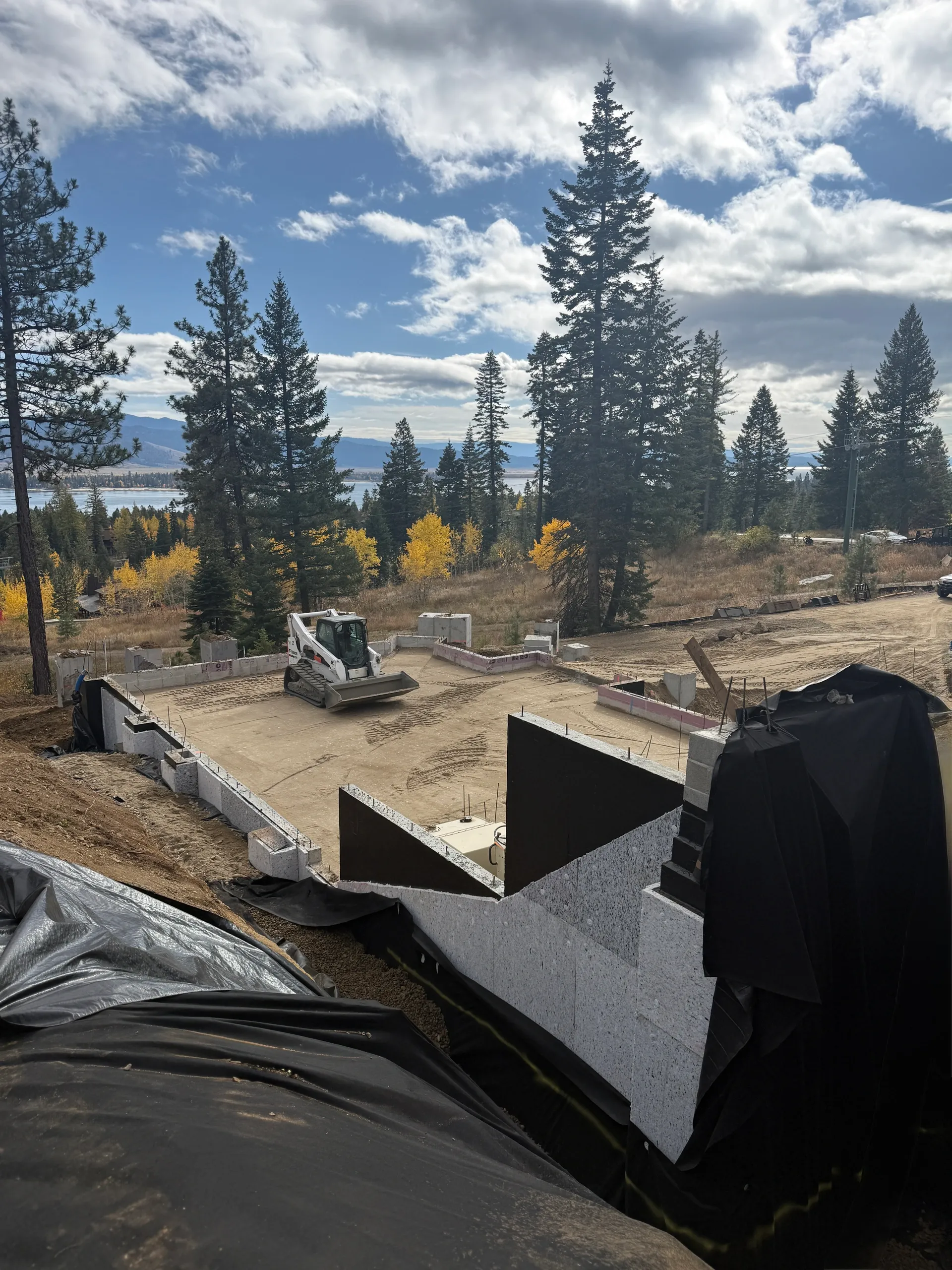 Construction site with excavator on a foundation, surrounded by tall pine trees, with a scenic lake and mountains in the background under partly cloudy sky.