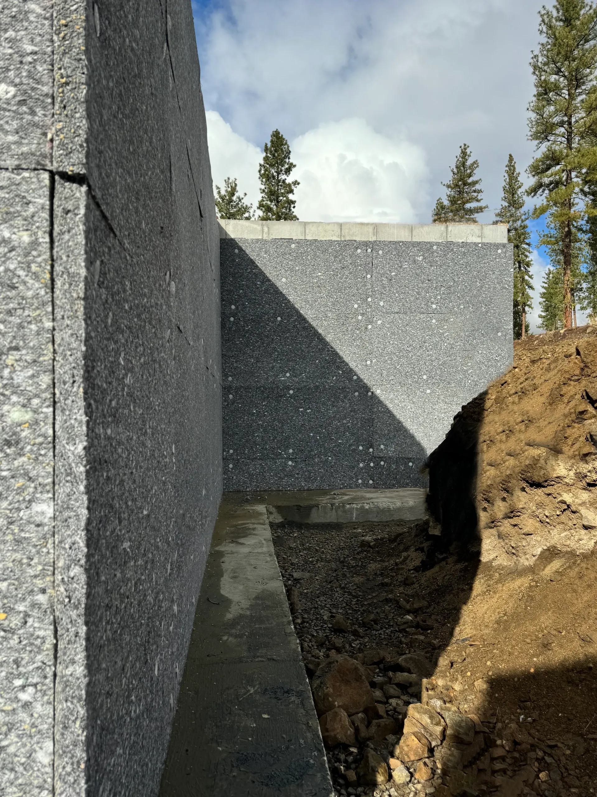 Construction site with gray concrete walls, dirt and rocks on the ground, and trees in the background, under a partly cloudy sky.