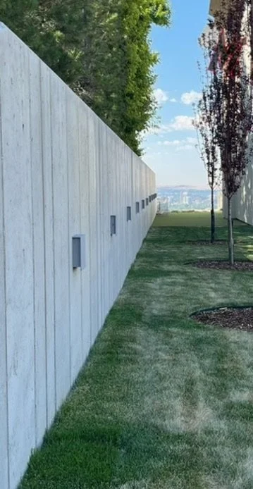 A concrete wall with small square windows, a grassy area, and trees with a city view in the distance under a partly cloudy sky.