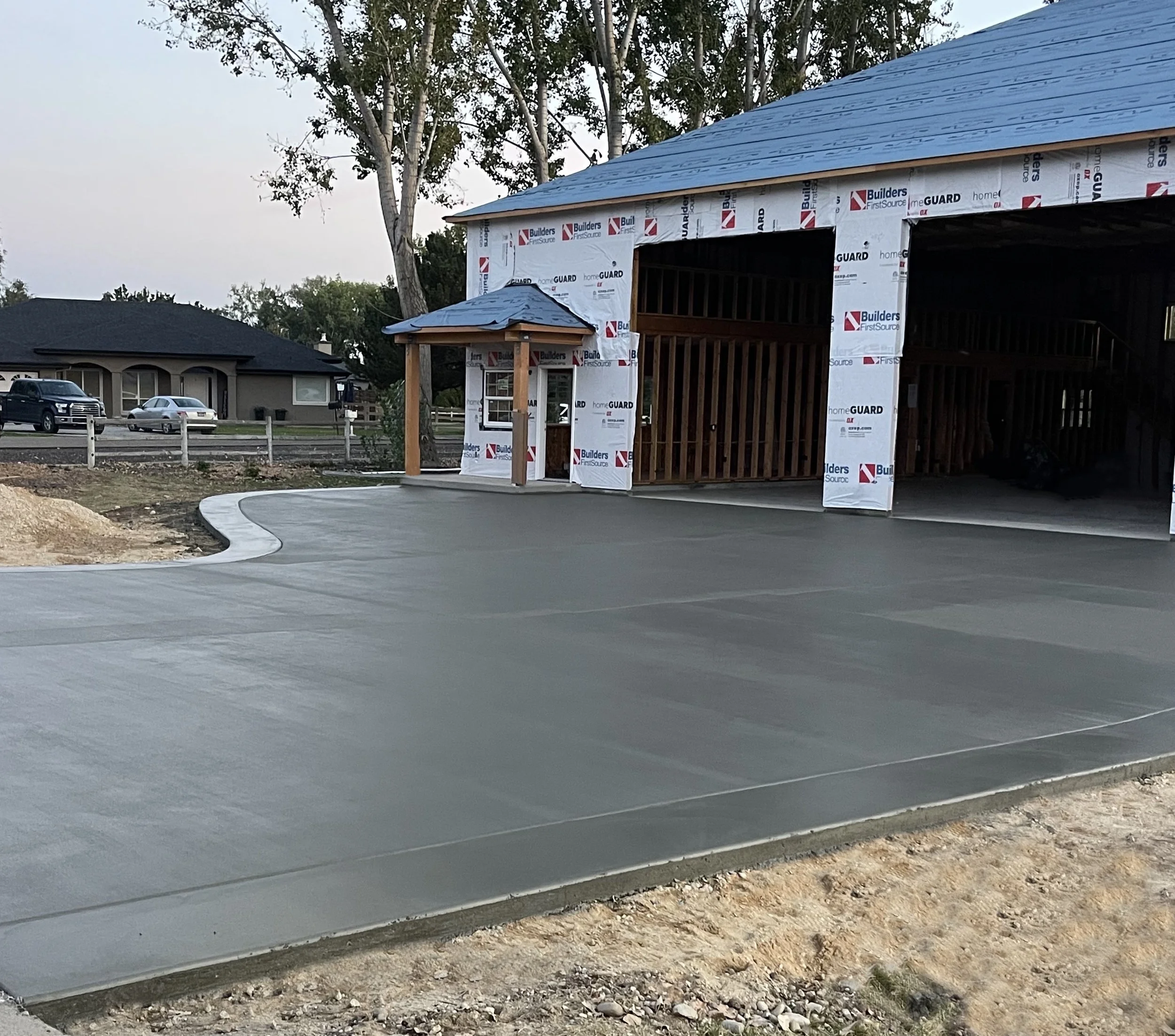 Under-construction garage or building with new concrete driveway, construction materials, and a small porch area with a blue roof, in a residential neighborhood.