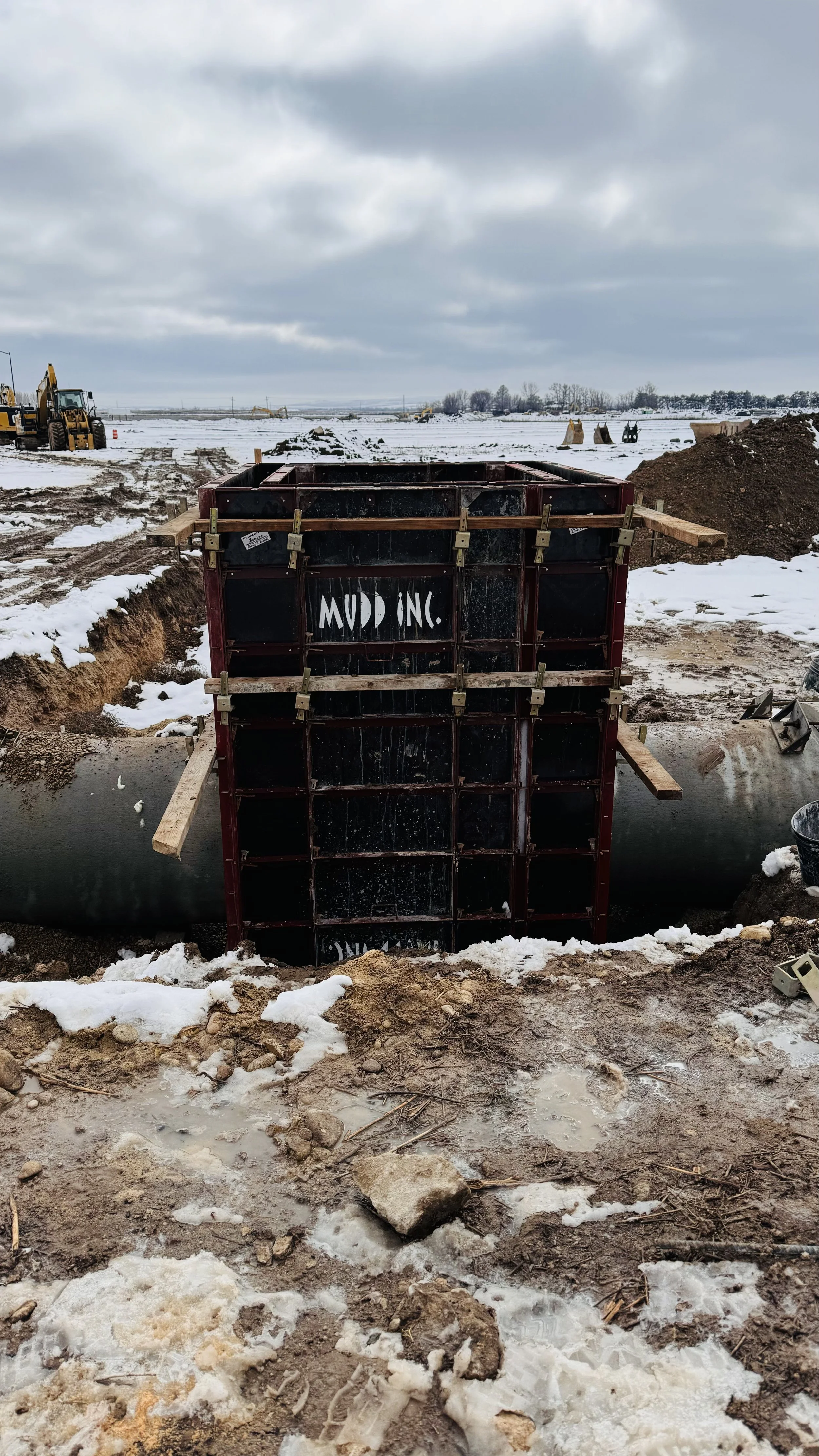 Construction site with large pipe and a concrete form with the words 'MUD INC' on it, snow-covered ground, with construction equipment and excavators in the background.