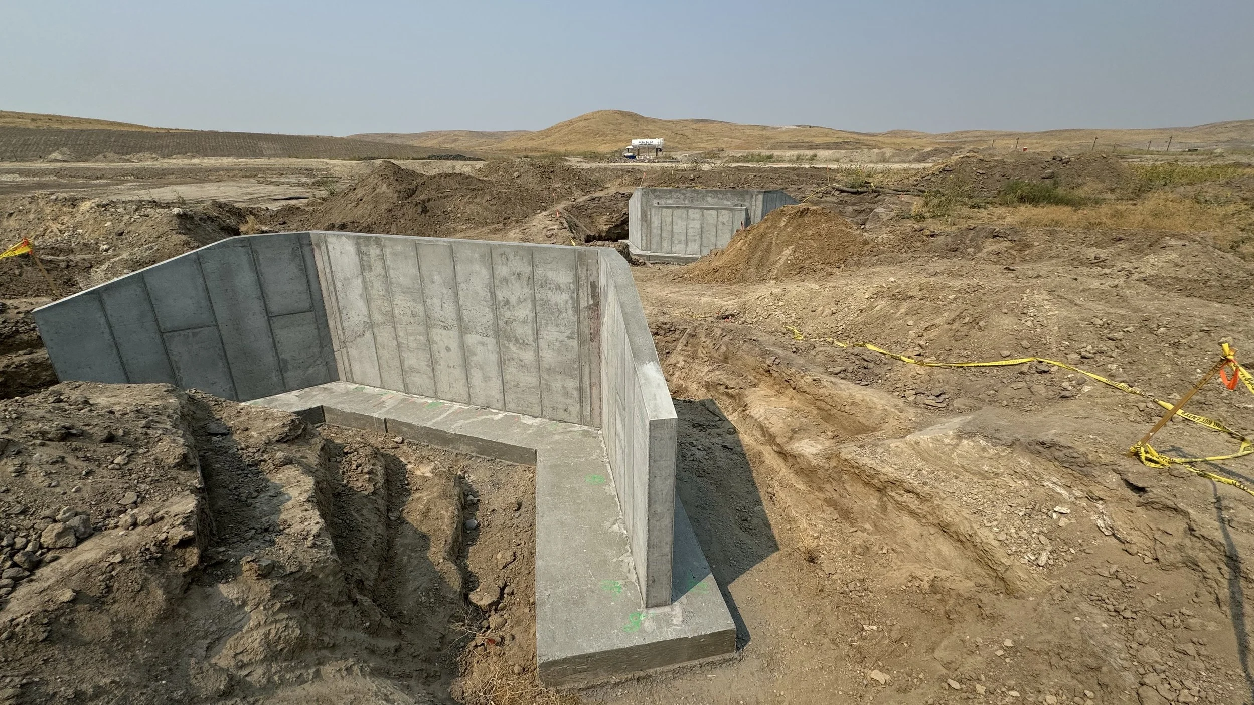 Construction site with large concrete barriers and dirt mounds in a rural area with hills in the distance.