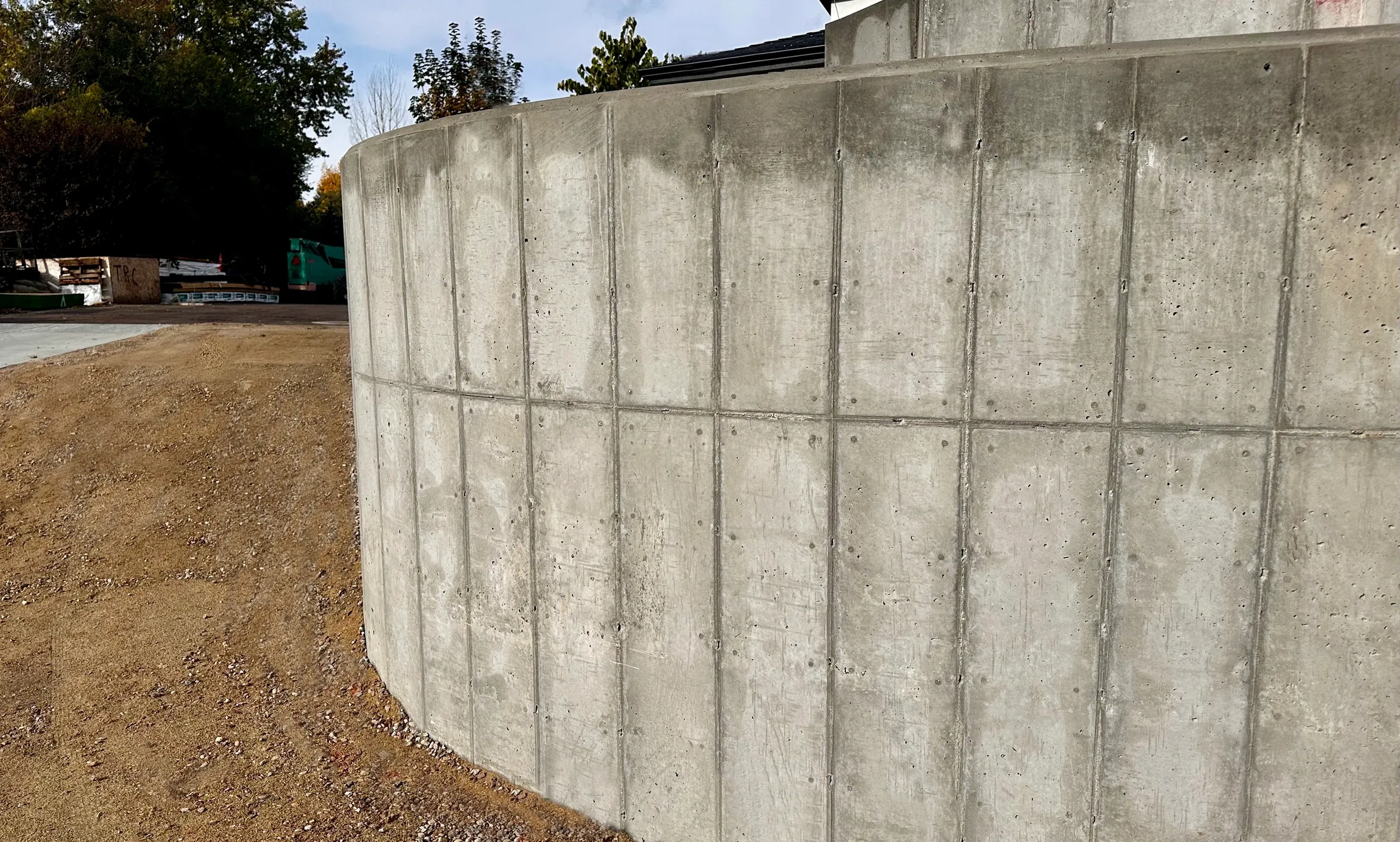A curved concrete retaining wall at a construction site with earth and gravel in the foreground and trees and a building in the background.
