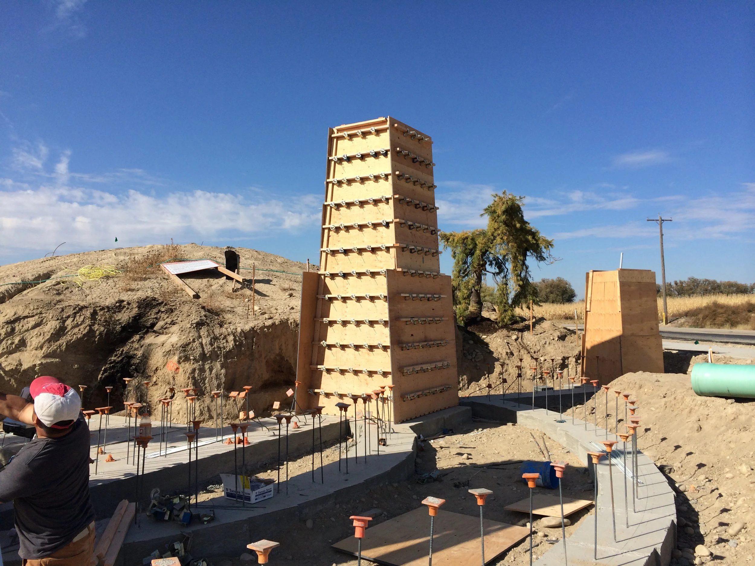 Construction site with wooden formwork for concrete, including a tall climbing wall, concrete foundation, metal rebar stakes, and a worker wearing a red and white cap in the foreground.