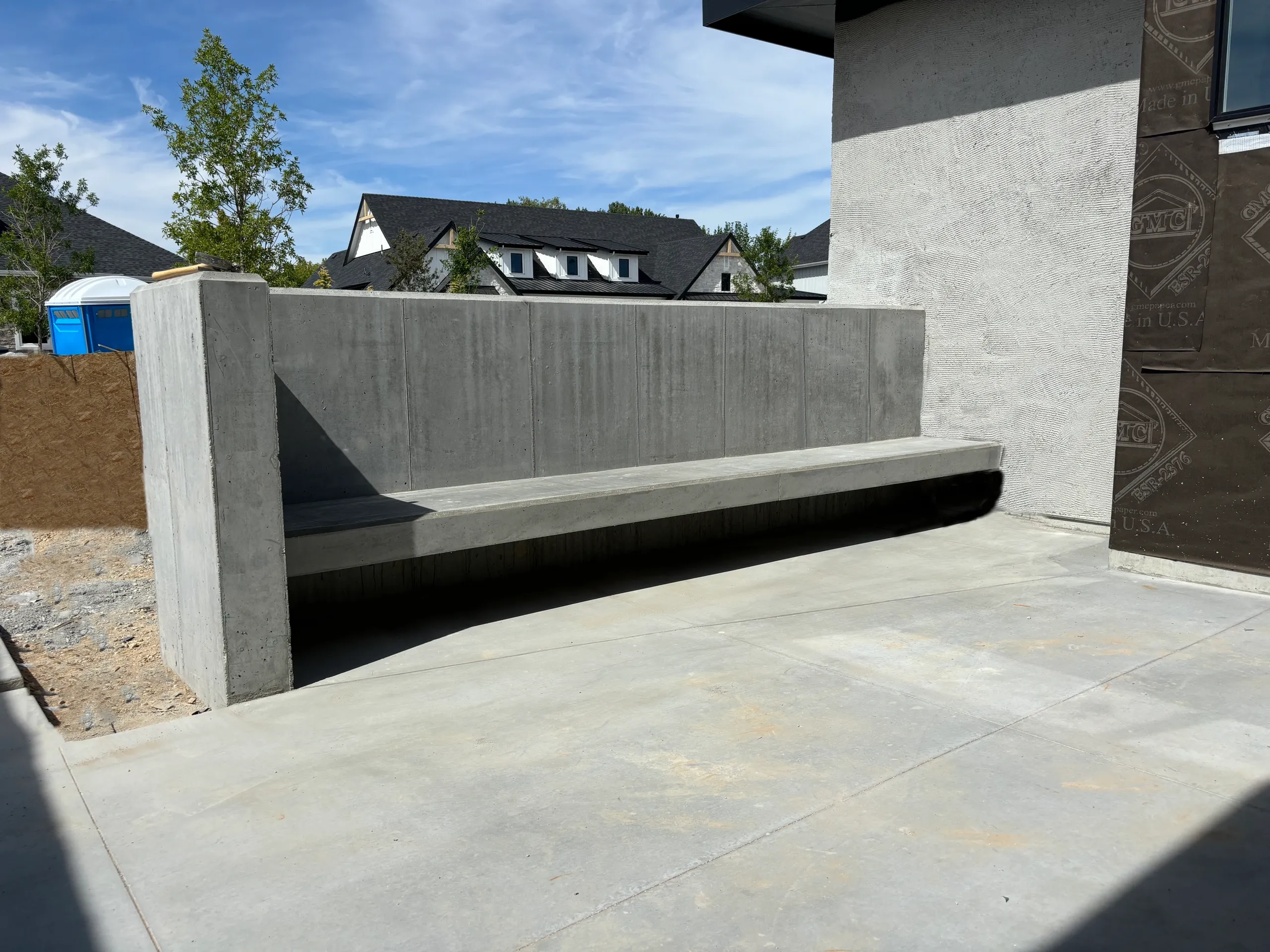 Concrete bench under construction on a paved area outside a building, with houses and trees in the background.