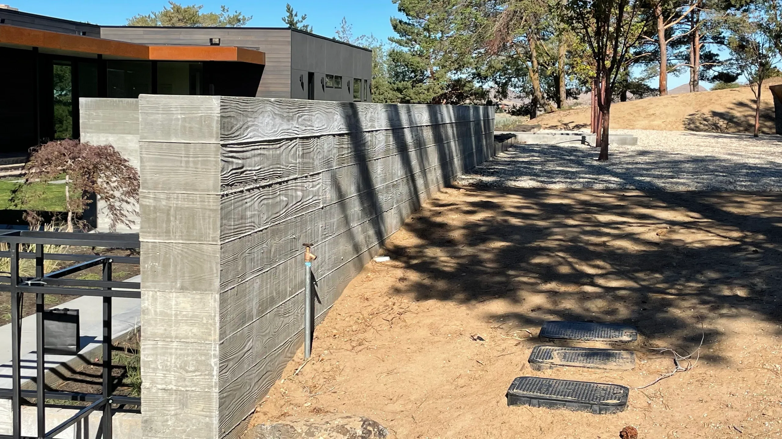 A modern yard with a tall wood privacy wall, trees, and a building in the background, with shadows of tree branches on the dirt ground, and three black landscape edging pieces on the ground.