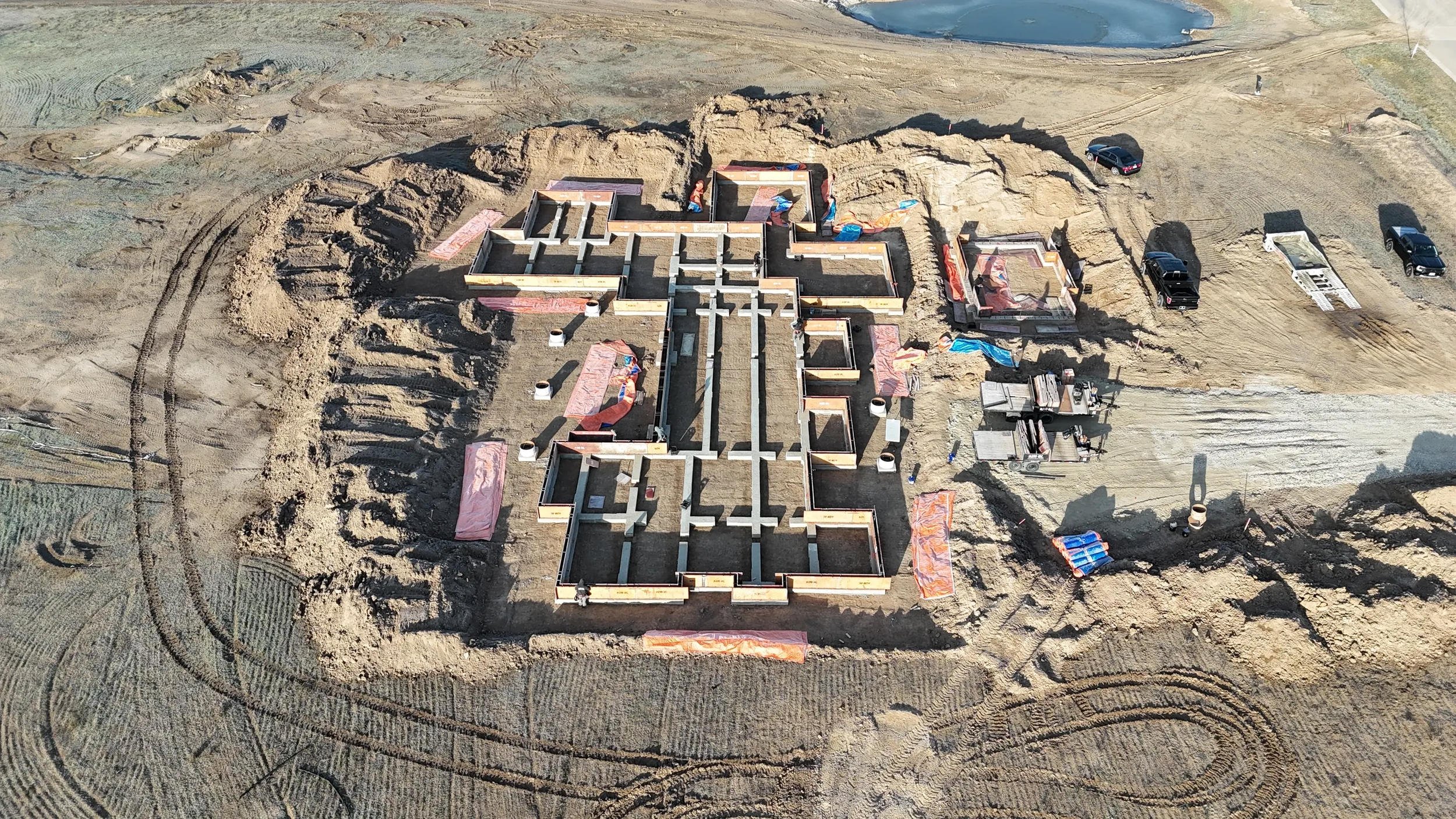 Aerial view of a construction site with the foundation of a building in shape of a cross, concrete walls being constructed, construction vehicles, and parked cars on dirt and gravel roads. Earth-moving tracks surround the structure.