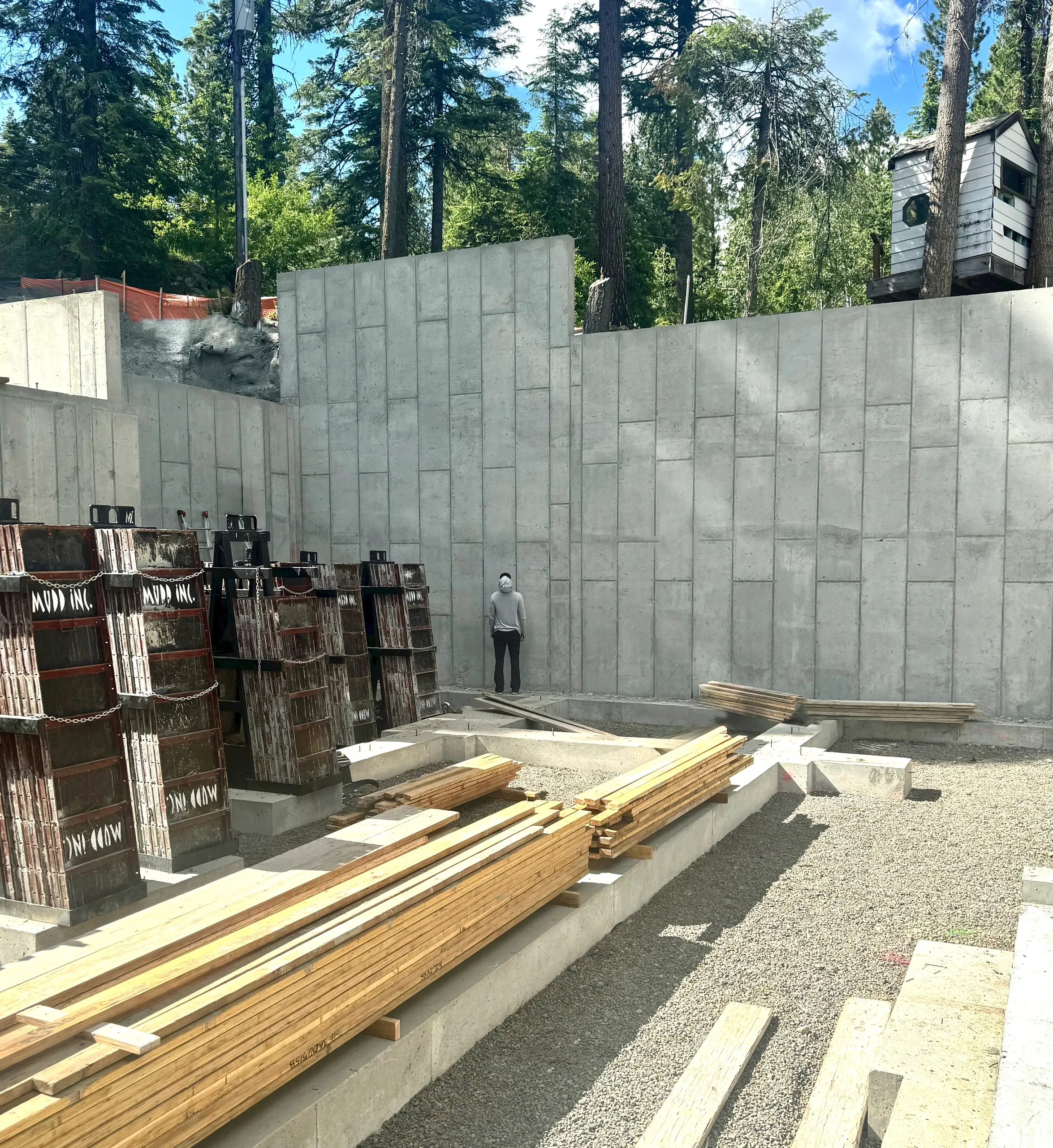 Construction site with a large concrete wall, wooden planks stacked, and construction equipment, with trees in the background.