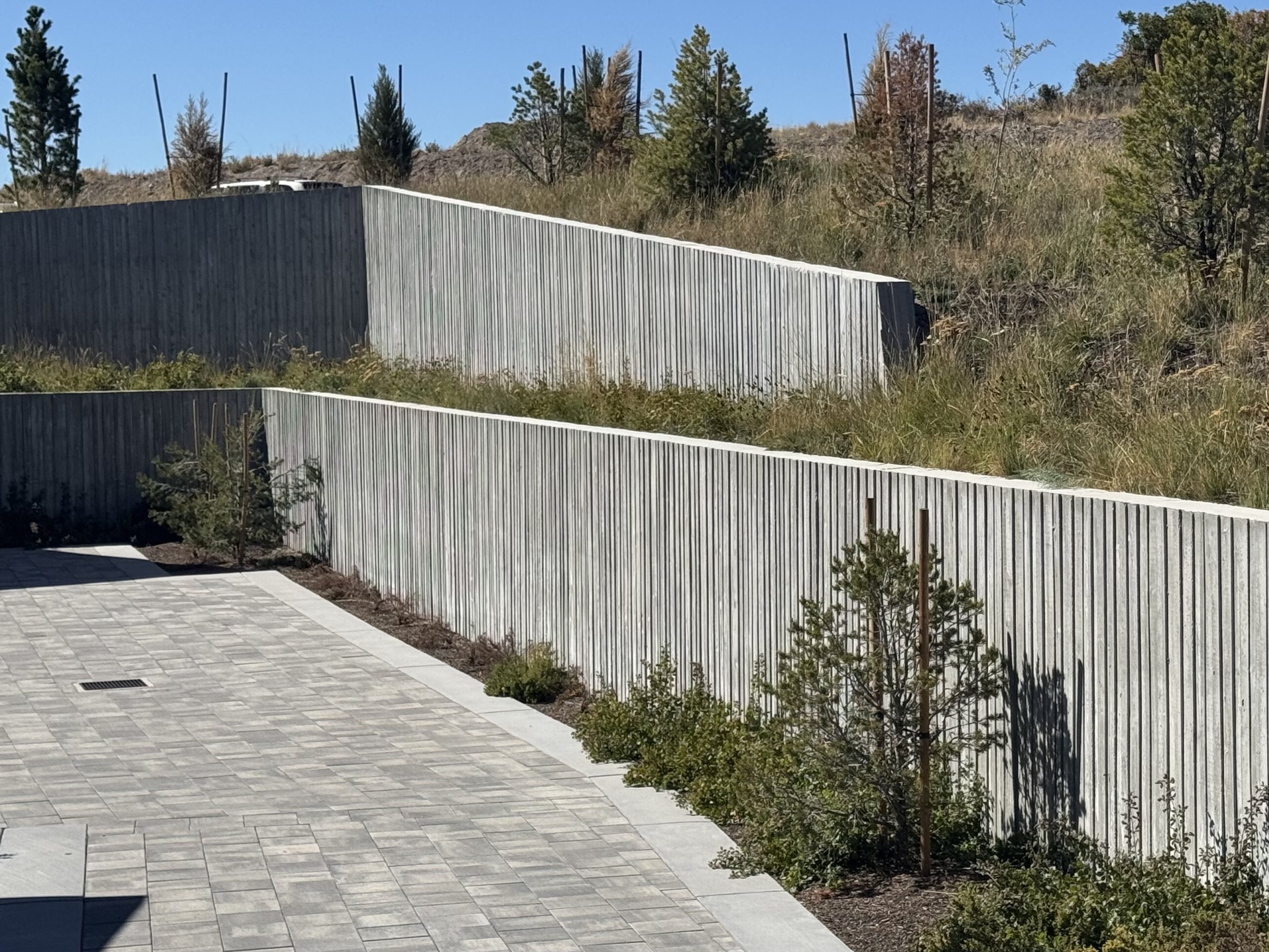 A concrete patio area with a small shrubbery and a landscapedled yard, enclosed by a tall wooden privacy fence with trees and a hillside in the background under a clear blue sky.