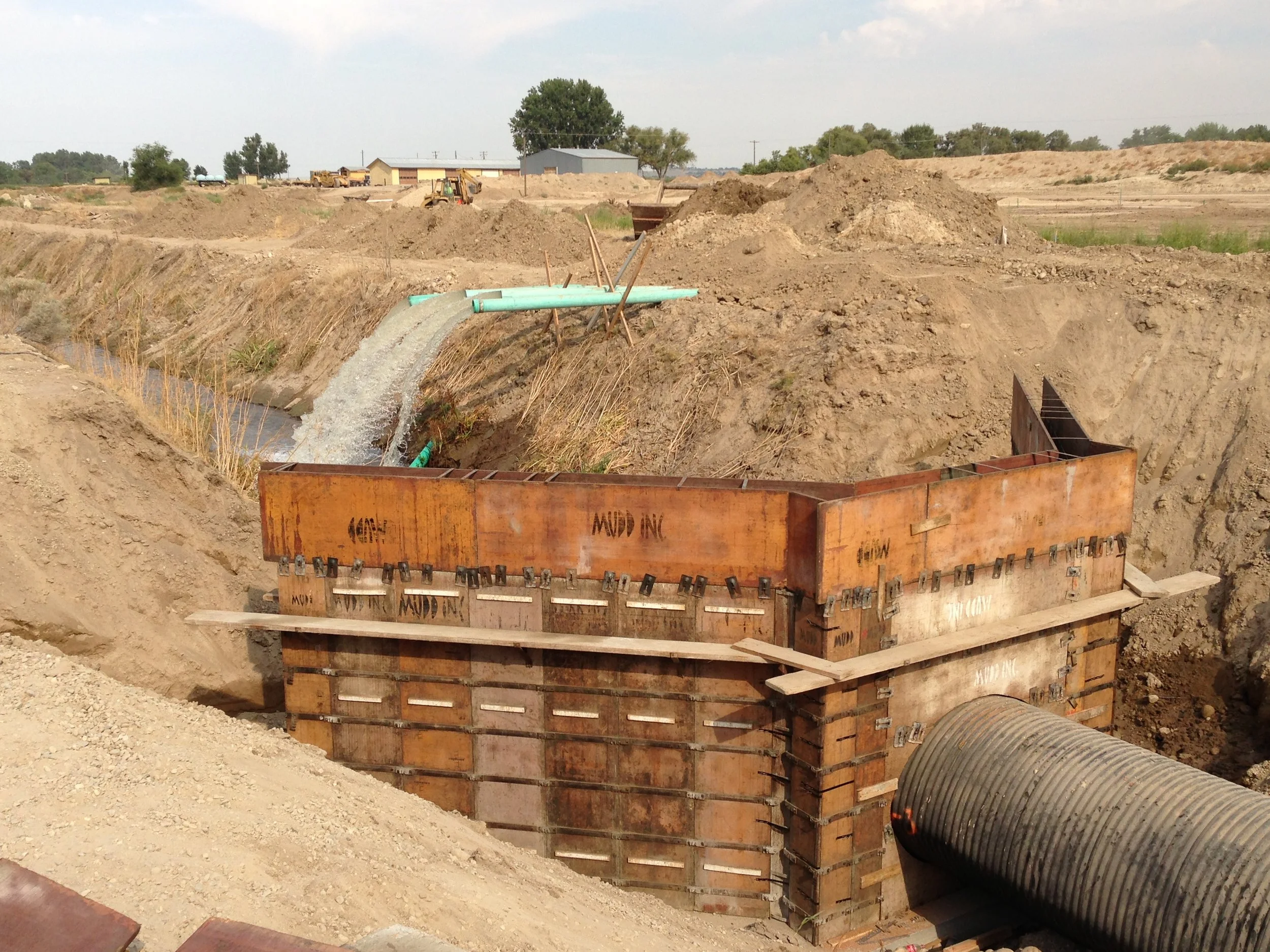 An ongoing construction project shows a large concrete form in a trench for installing a pipeline, with pipes and construction equipment visible in the background.