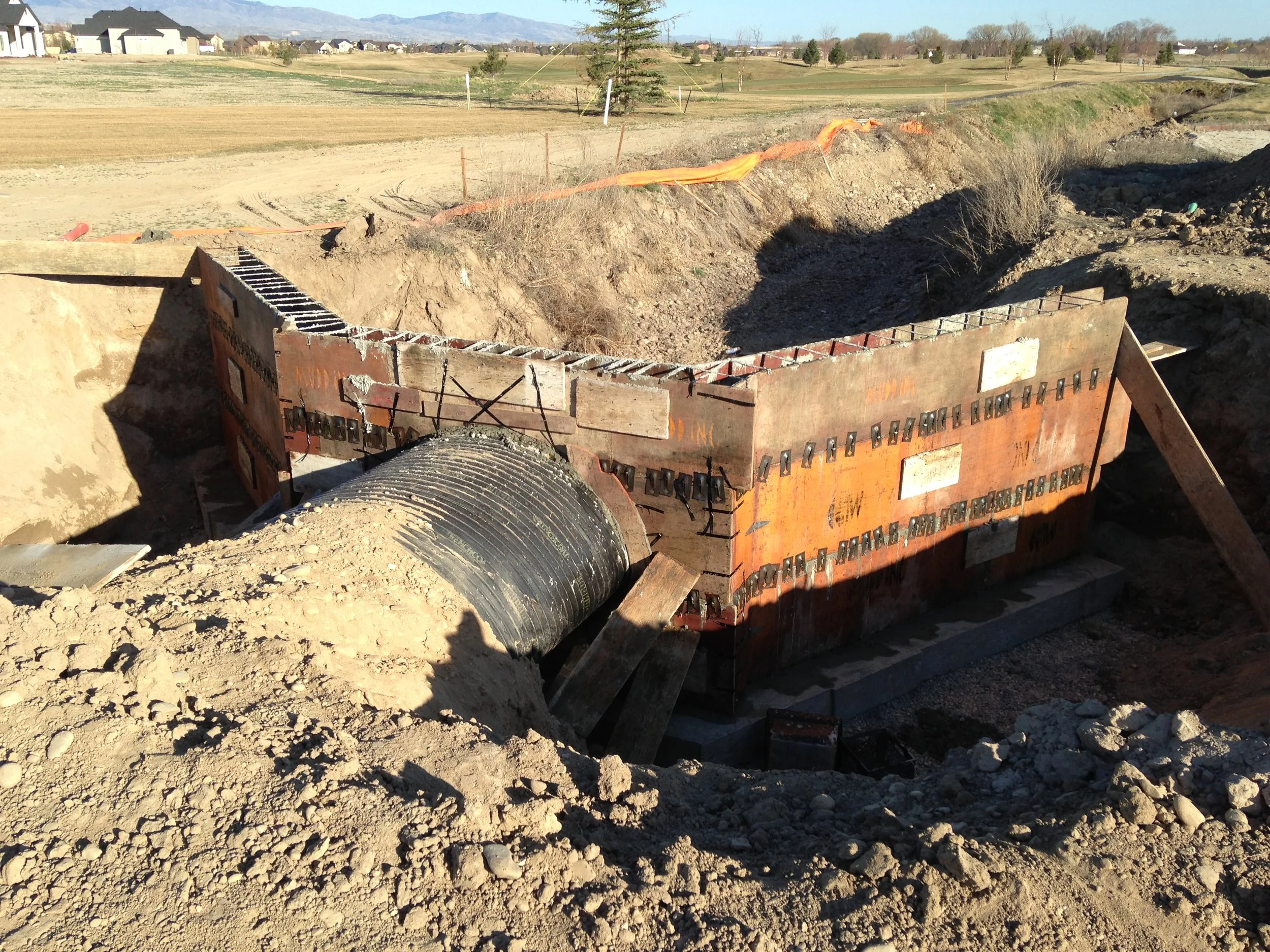 Construction site with a large underground pipe connecting to a concrete foundation, surrounded by dirt and soil, with a rural landscape in the background.