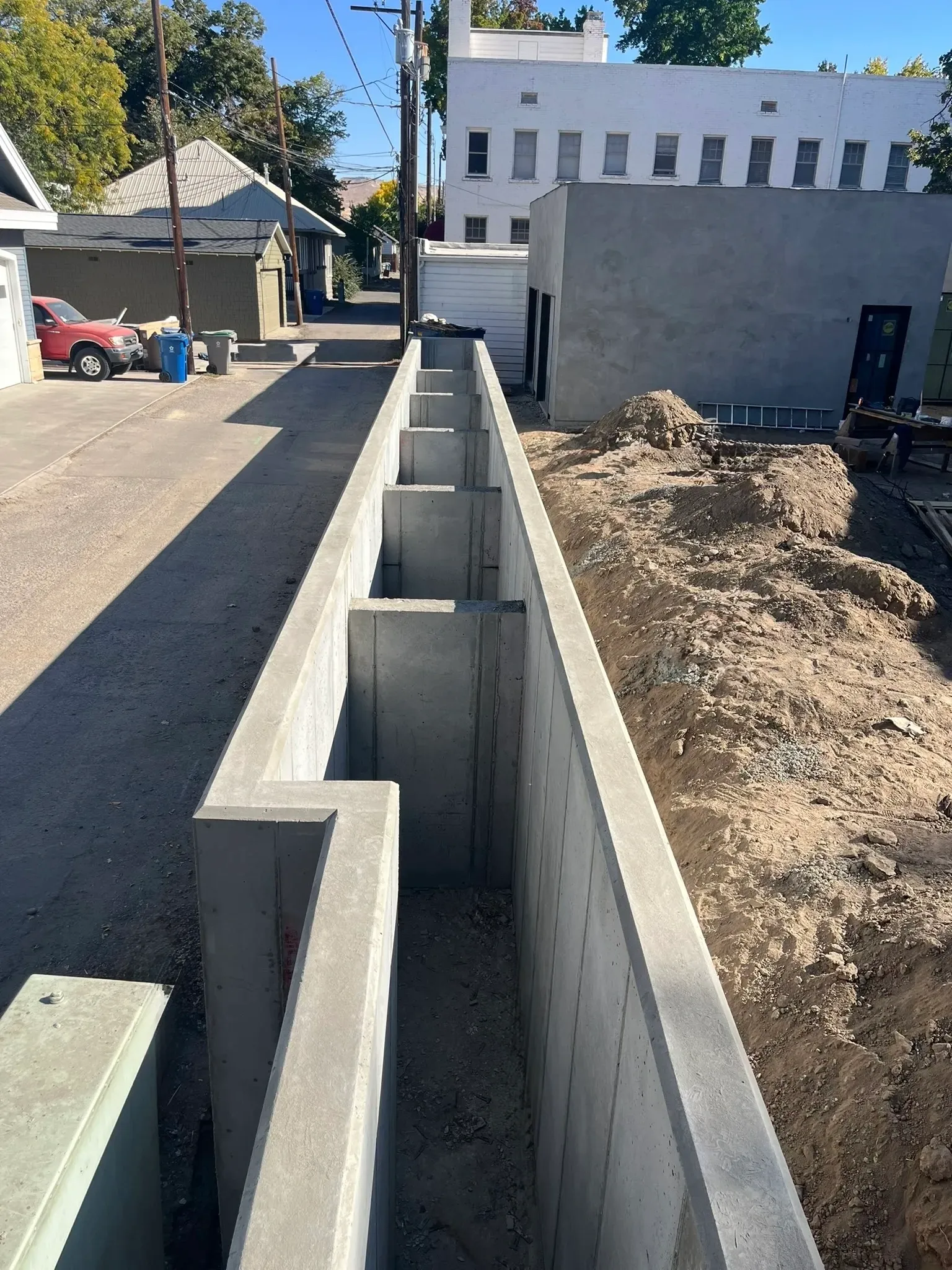 Concrete foundation blocks are being installed along a construction site in a residential neighborhood. There are houses and trees in the background, with some construction equipment and materials nearby.