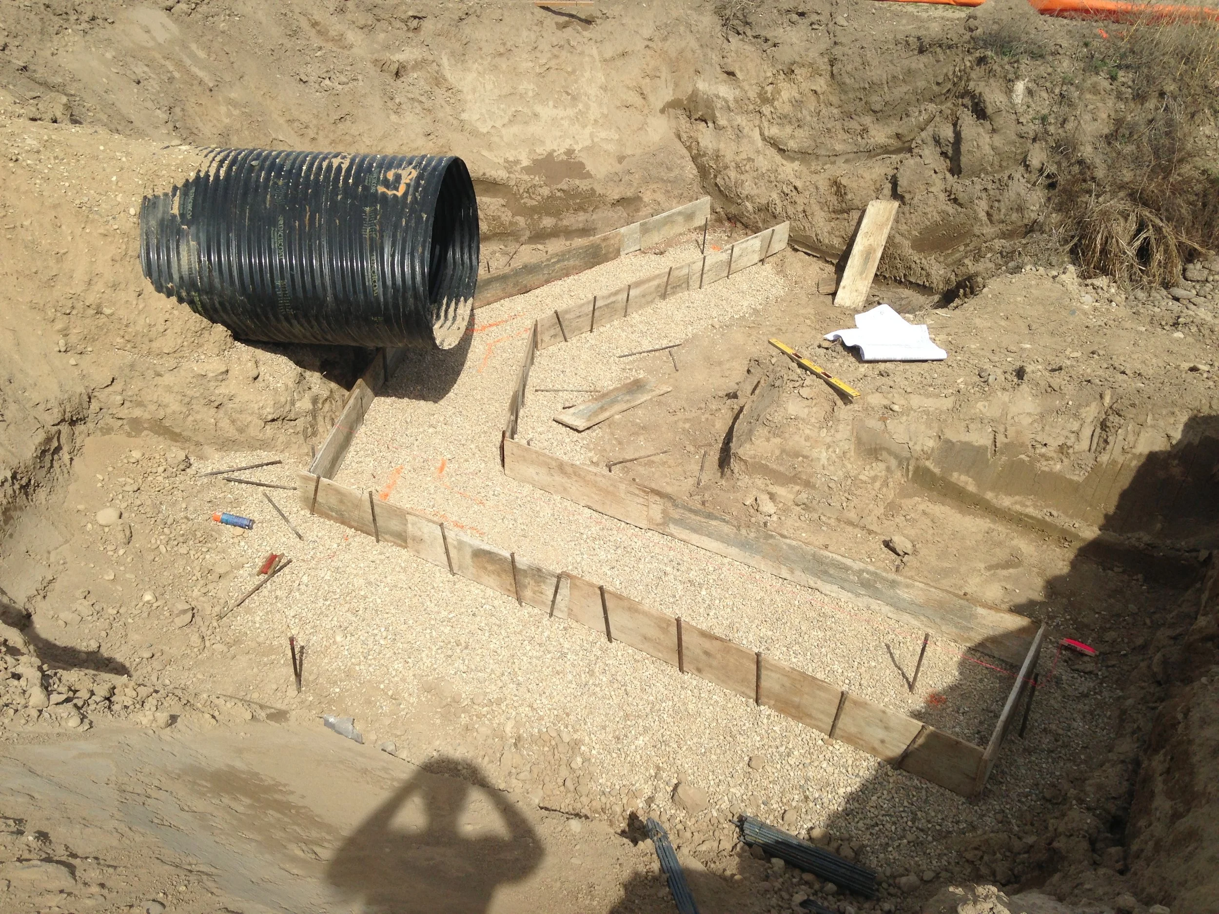 Construction site excavation with wooden and metal framing, gravel, and a large black pipe, likely for a foundation or drainage system.
