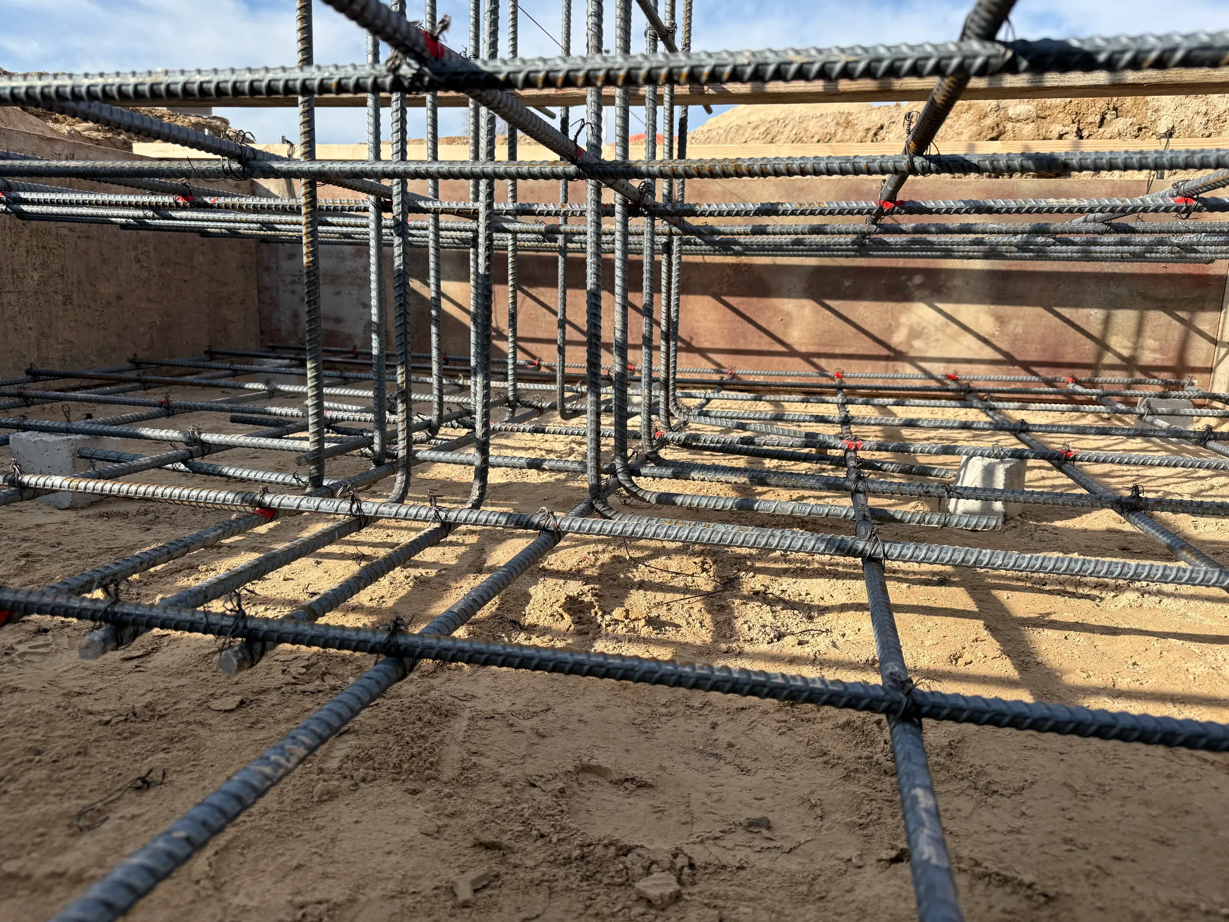Close-up of steel rebar grid framework at a construction site, set in sandy ground, with wooden formwork in the background, under a clear sky.