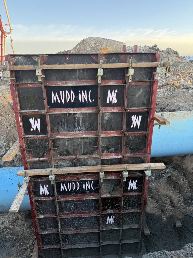 Construction site with a large metal formwork for concrete, supported by wooden beams, and a blue pipe in the background. The formwork has logos and text reading 'Mudd Inc' and other symbols.