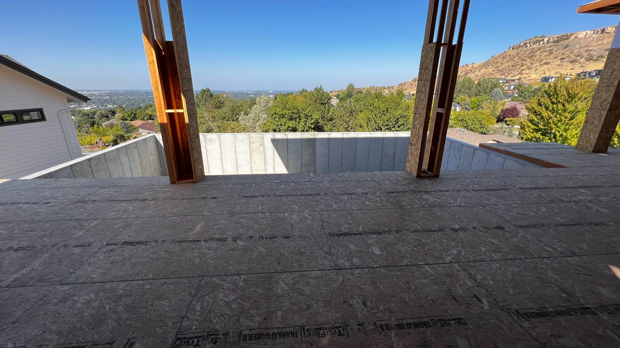 View from inside a building under construction, showing unfinished flooring and framing, overlooking a suburban neighborhood and distant hills under a clear blue sky.