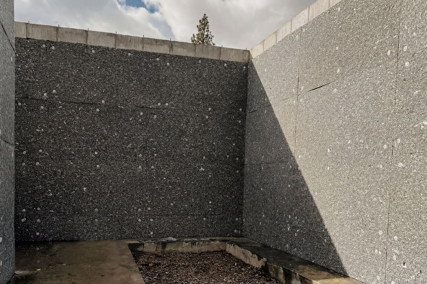 Concrete walls forming a corner with a patch of dirt on the ground, outside with cloudy sky visible overhead.
