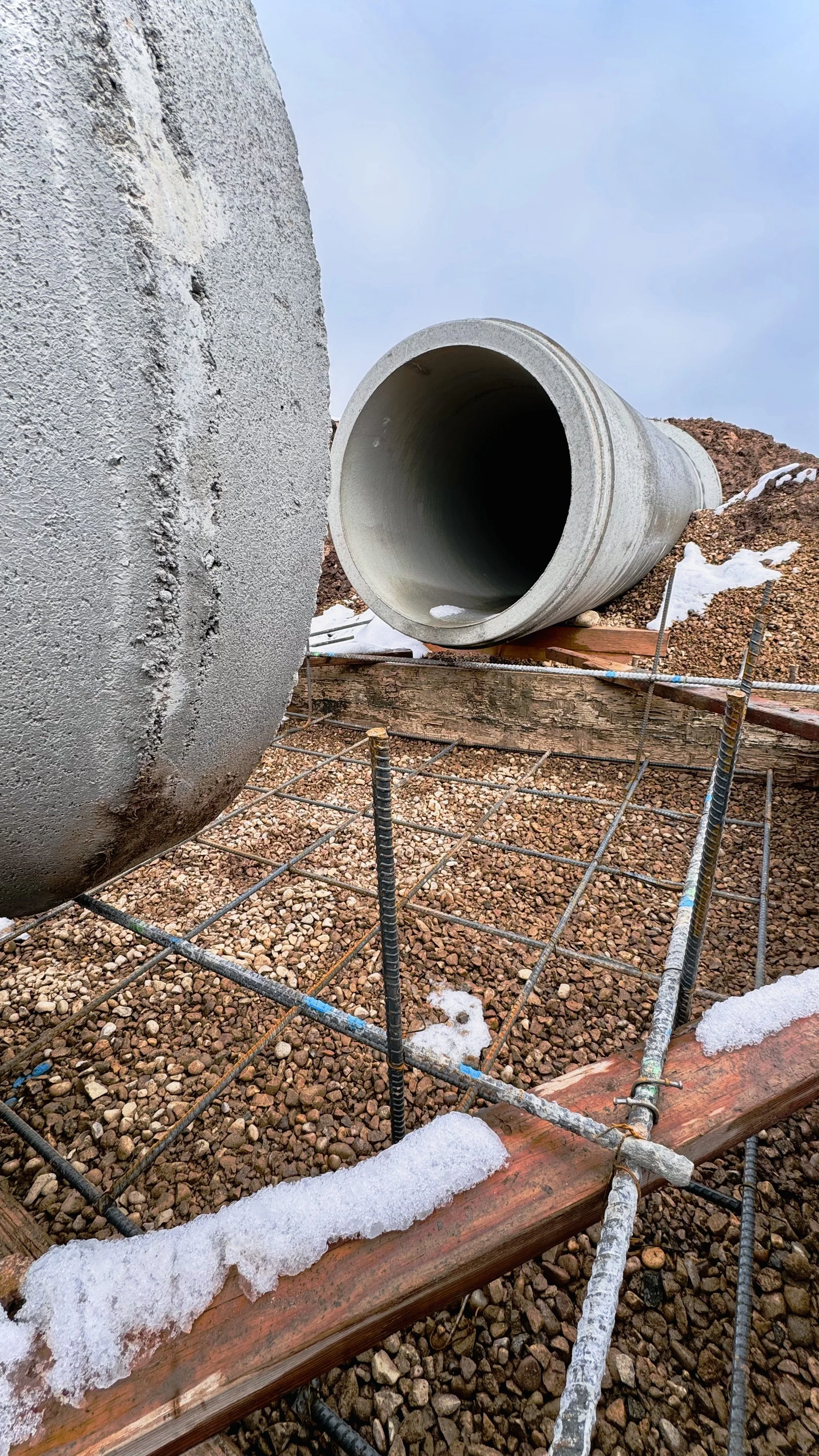 Close-up of a construction site with a large concrete pipe lying on gravel, surrounded by rebar and wooden forms, with snow patches on the ground and a cloudy sky.