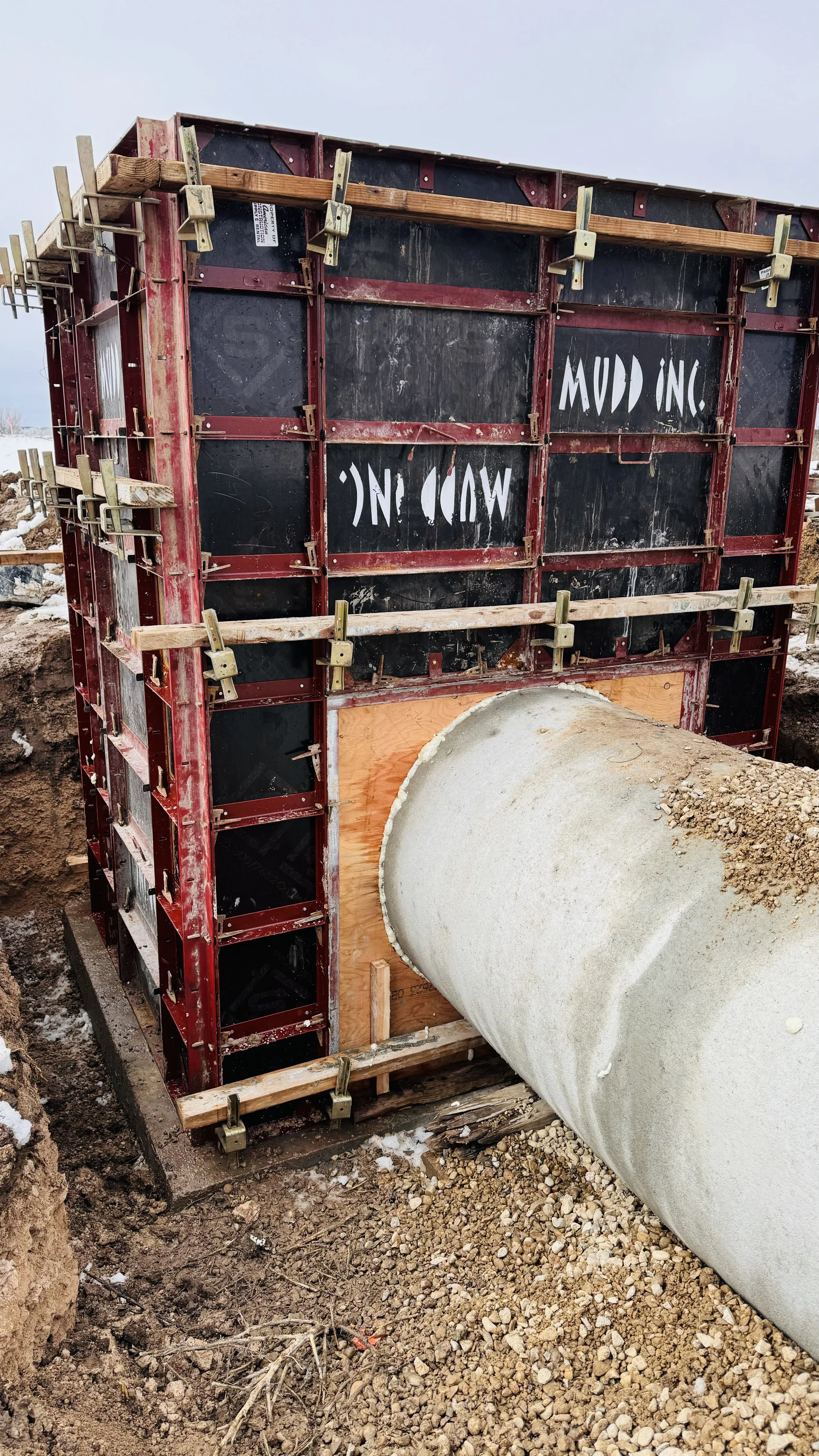 Construction site with a large concrete pipe being installed through an excavated section of earth, with formwork and framing for a concrete wall.