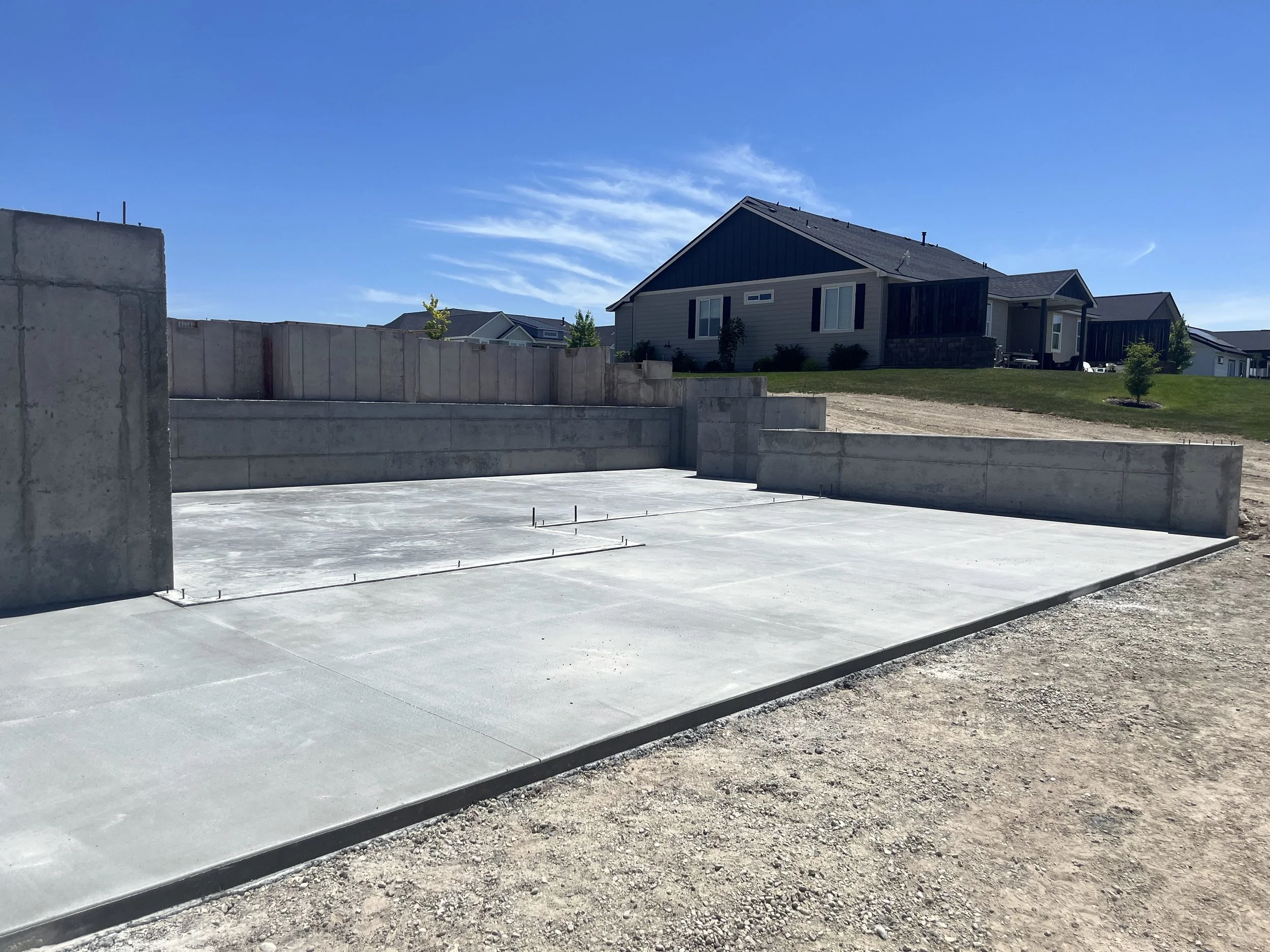 Concrete foundation structure of a building under construction with a clear blue sky in background and surrounding houses in a neighborhood.