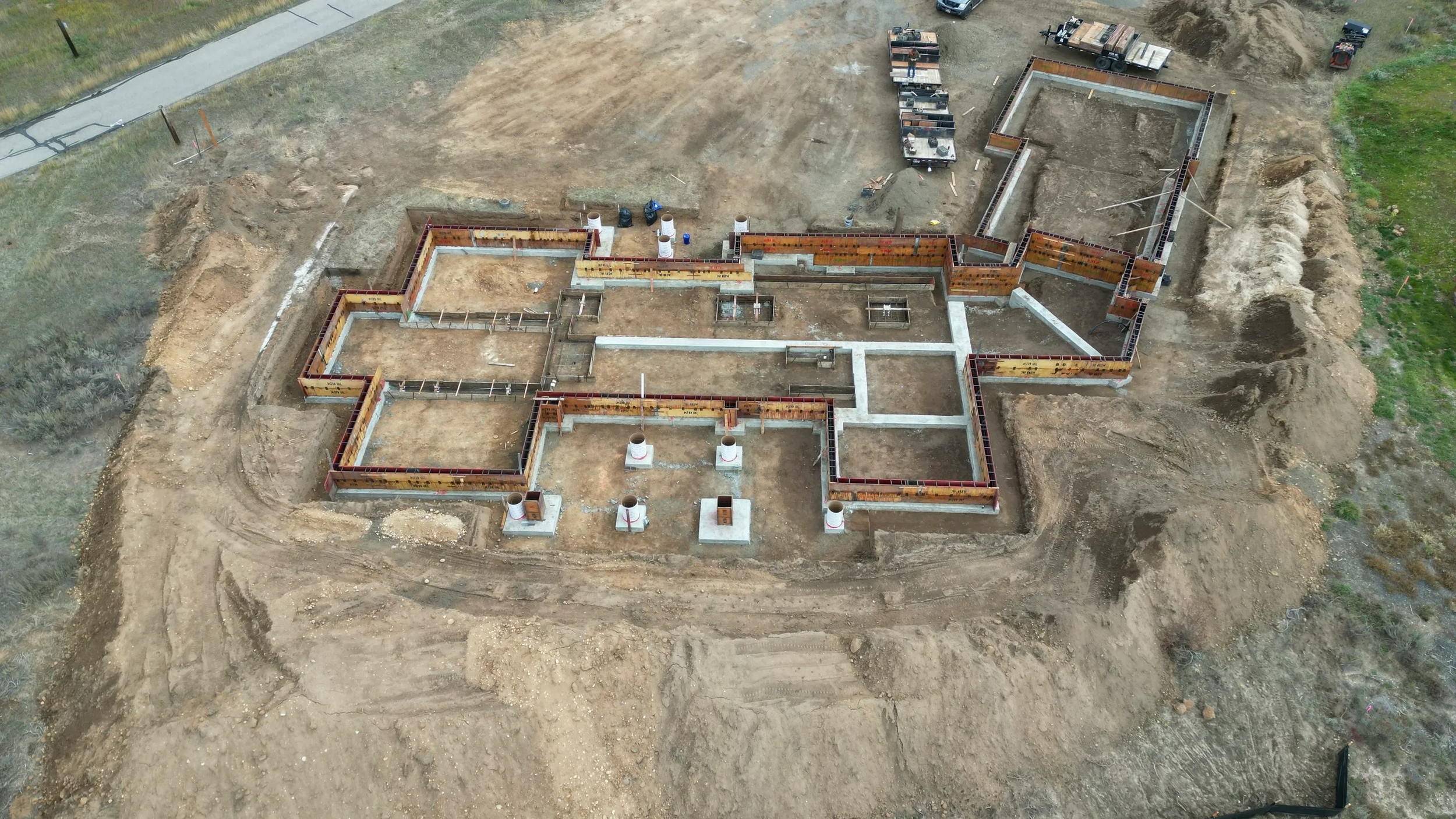 An aerial view of a house foundation under construction, showing concrete footings, framing, and plumbing pipes on a dirt lot.