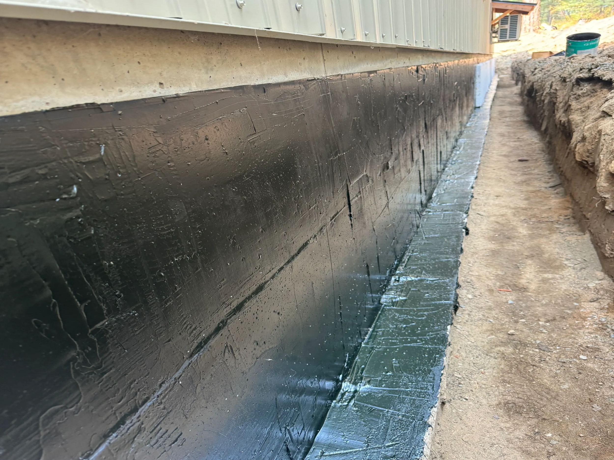 Close-up view of the foundation of a building with black waterproof coating being applied, with exposed dirt and gravel around.