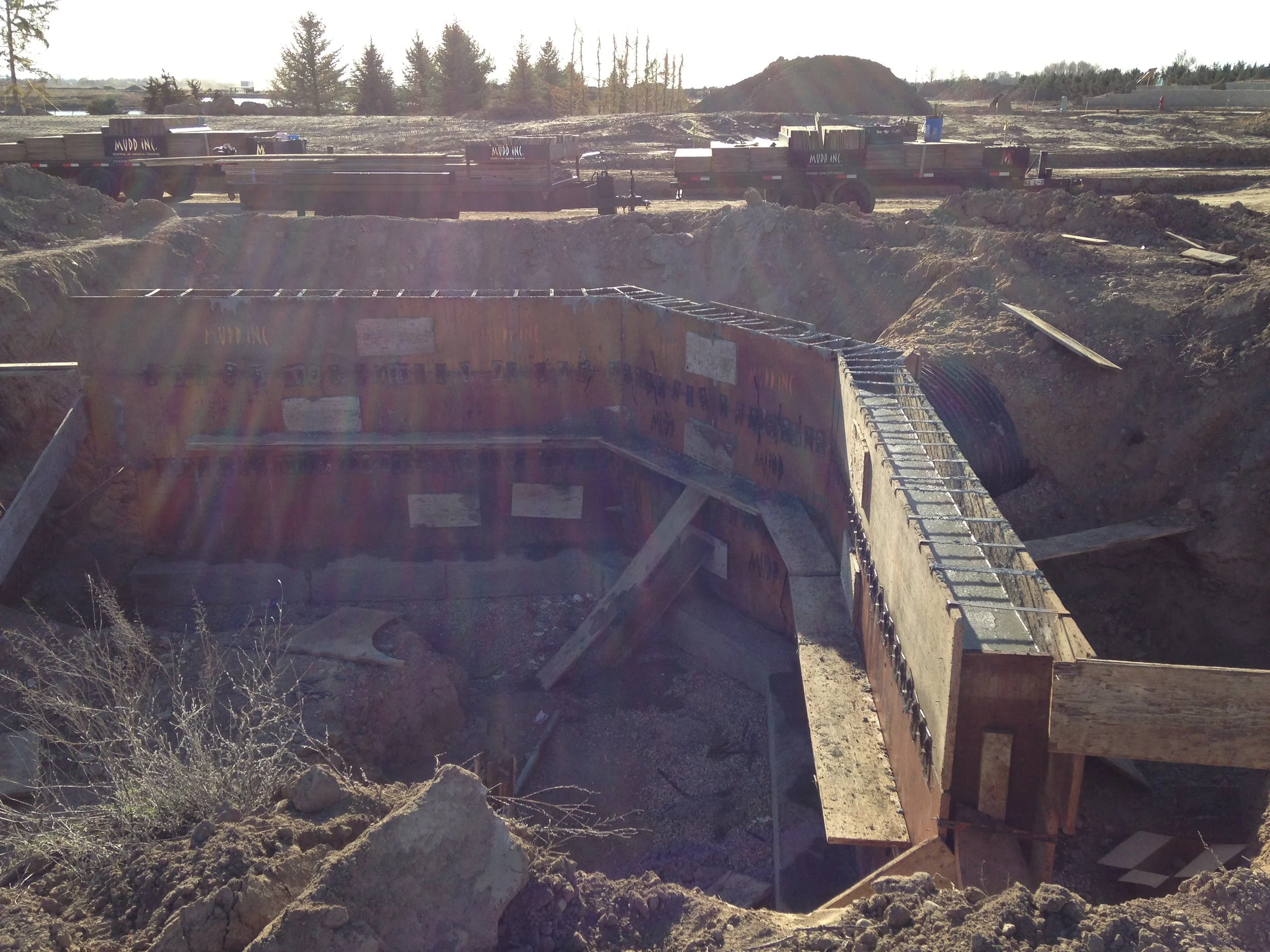 Construction site with a deep excavated hole for building foundation, with wooden and metal reinforcements and construction vehicles in the background.