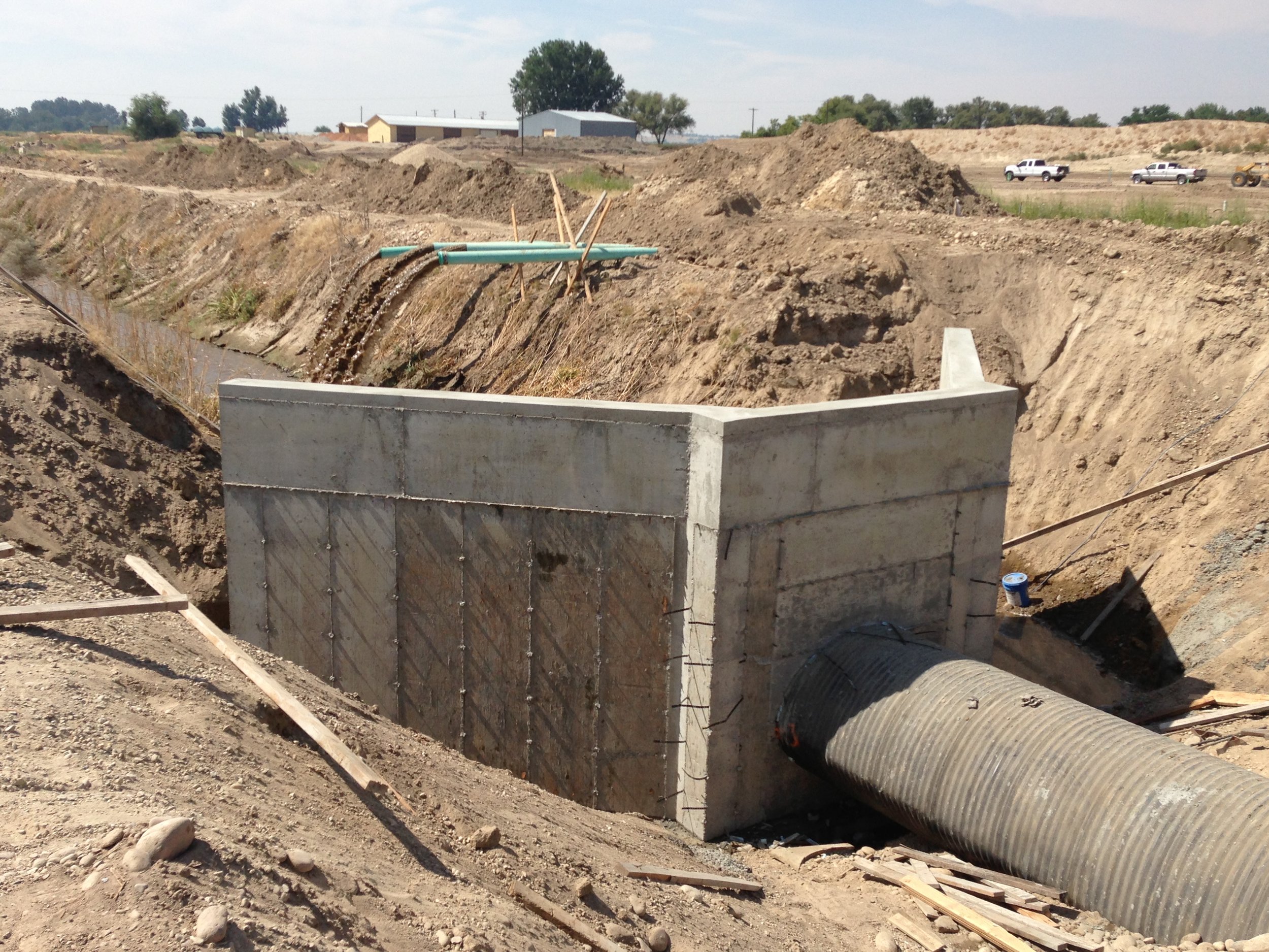 Construction site showing a concrete structure with a large pipe connected to a drainage system, surrounded by dirt and construction materials.