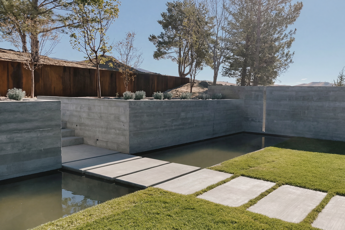 Minimalist outdoor backyard with concrete walls, stairway, small pond, well-maintained grass, and trees under a clear blue sky.