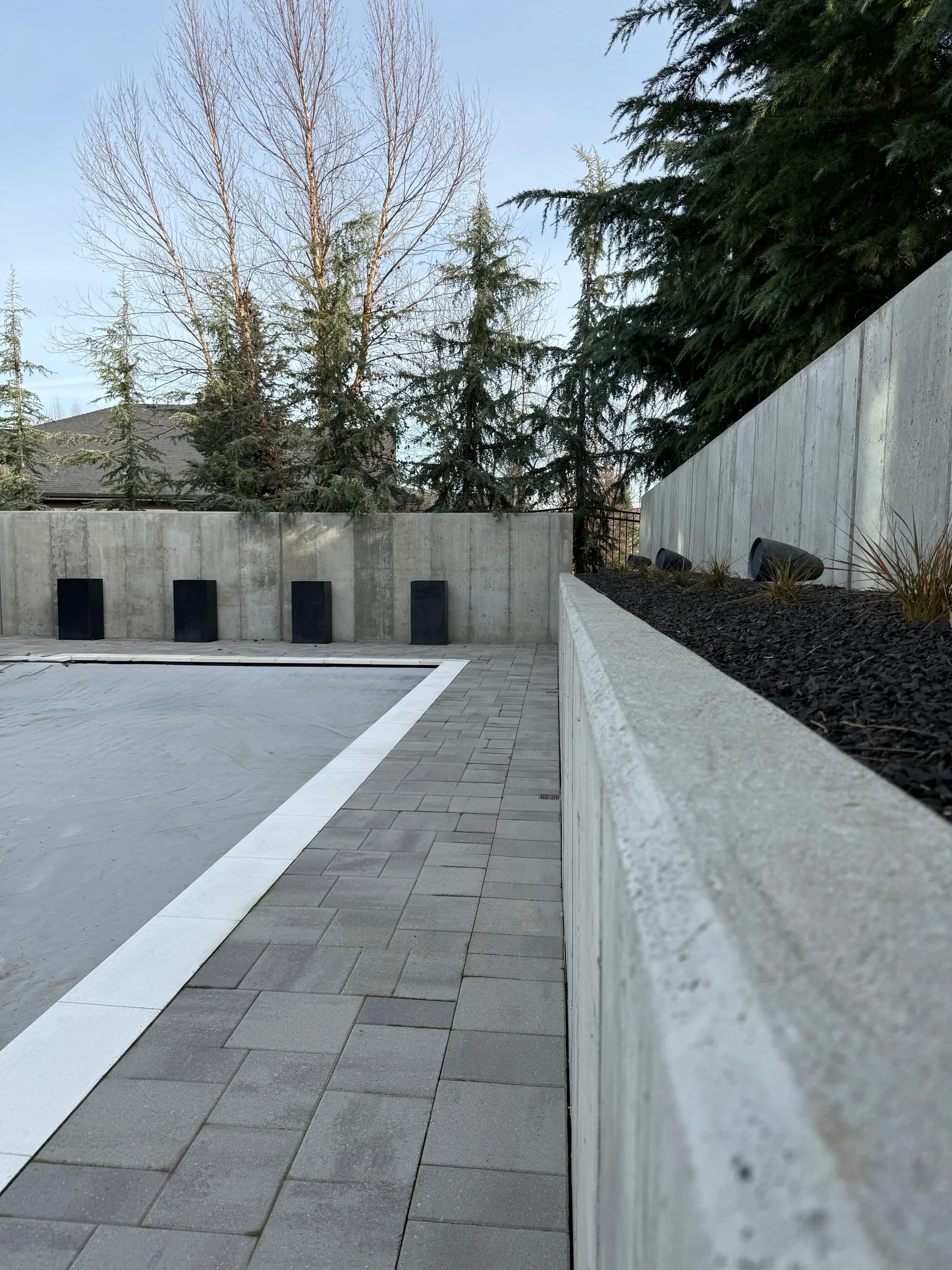Concrete wall with black planters on top, paved pathway with a white border, bordering a gravel area with ornamental plants, and evergreen trees in the background.