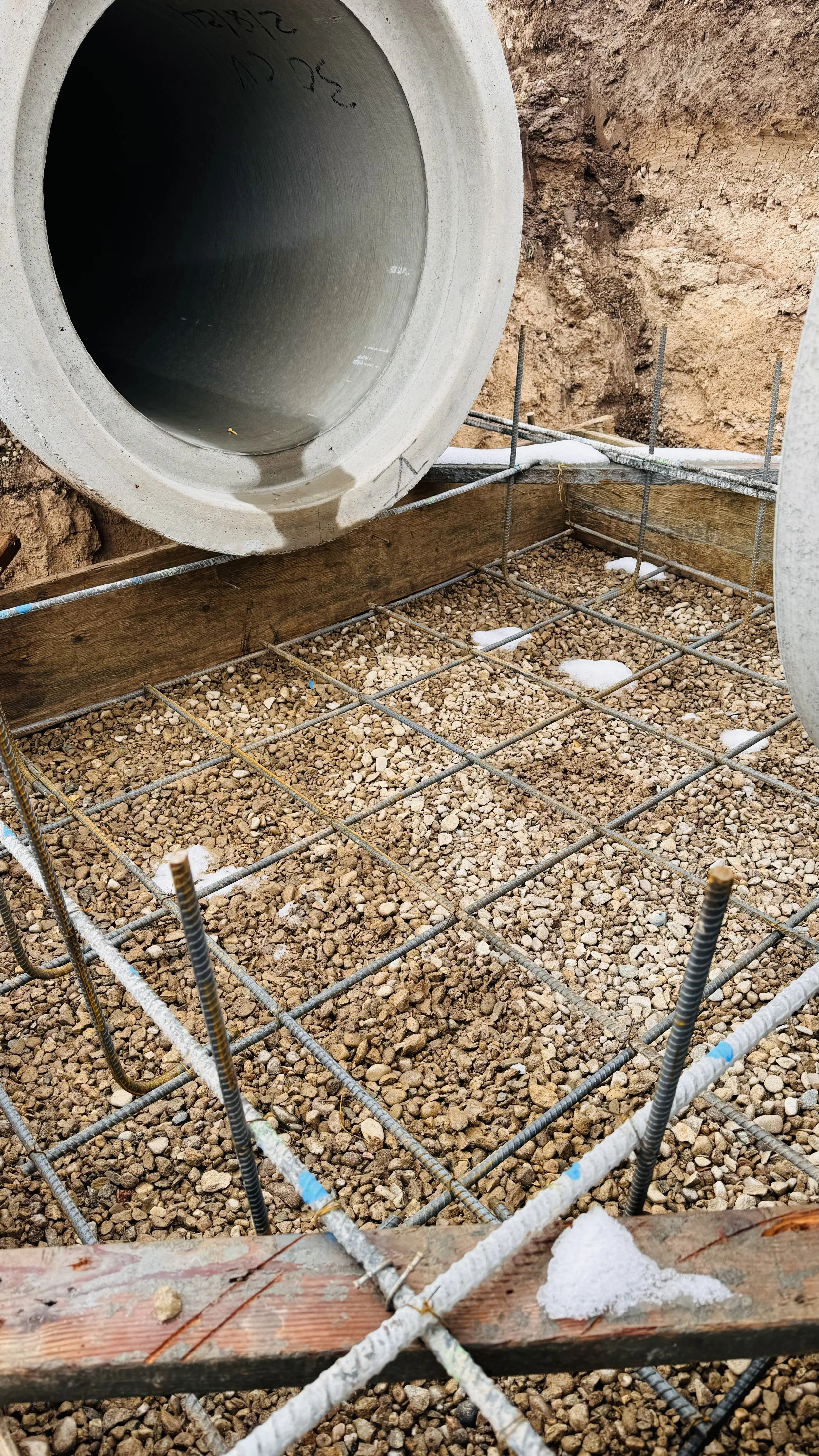 Construction site showing a concrete pipe, metal rebar, wooden formwork, and gravel ground.
