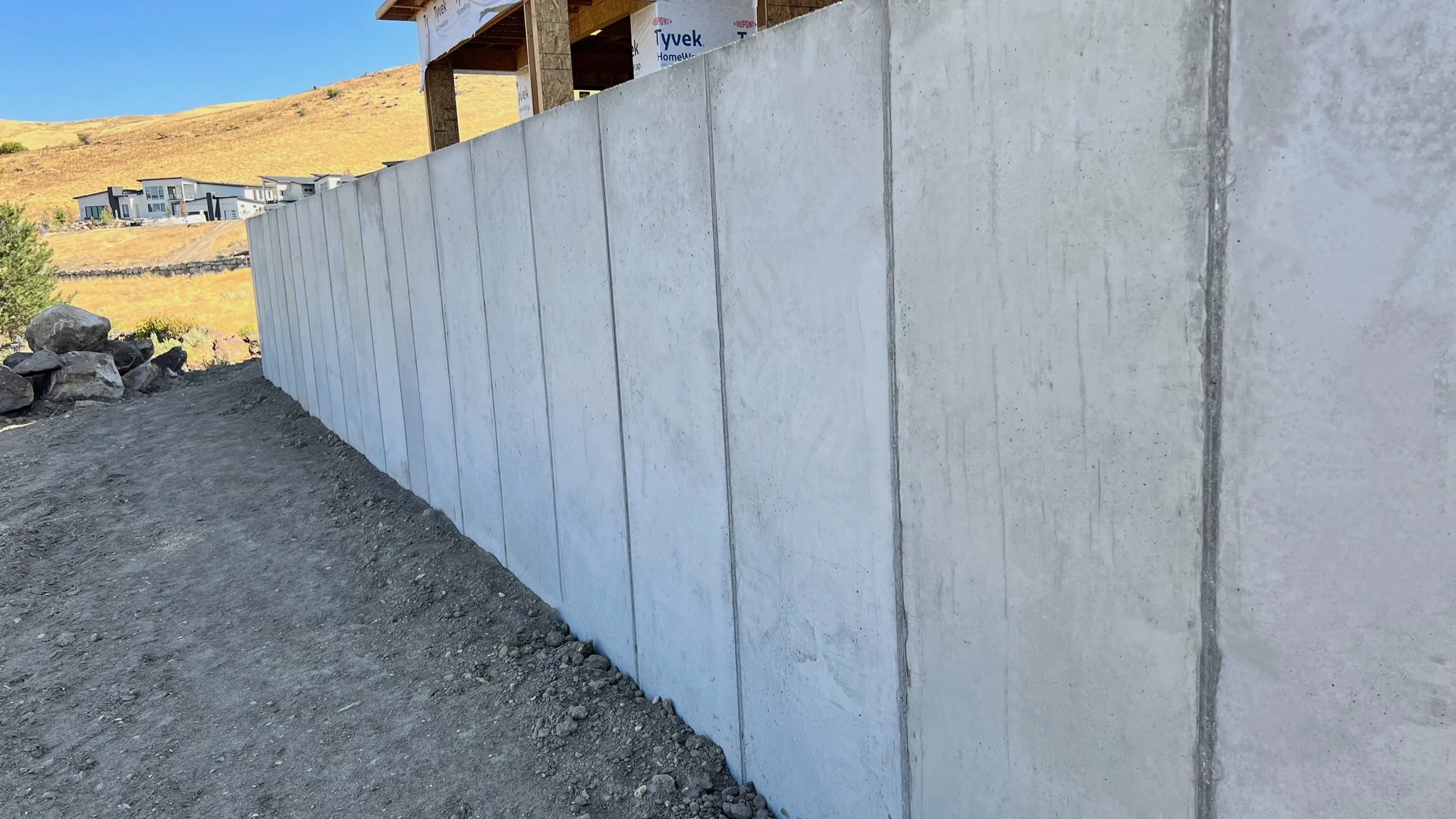 Concrete wall under construction on a sloped dirt ground at a building site, with houses and a hill visible in the background under a blue sky.