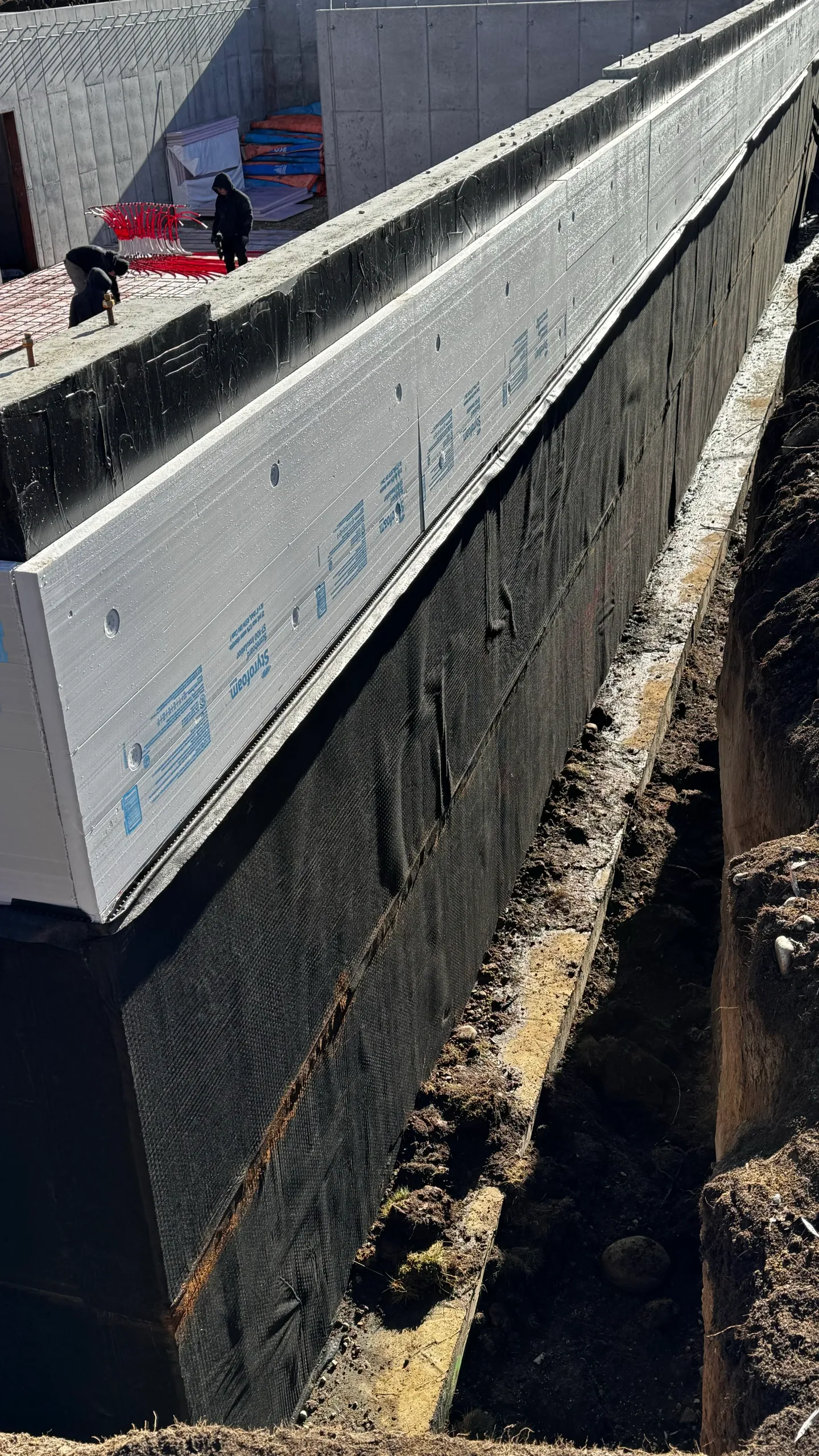 Construction site with workers installing insulation on a building's exterior wall, with construction materials and equipment visible in the background.