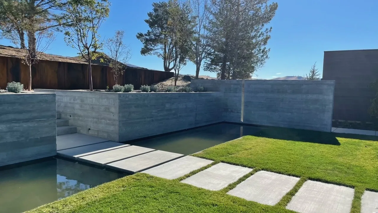 Modern backyard with concrete planters, a small bridge over water, a grassy area with stepping stones, and trees in the background under a clear blue sky.