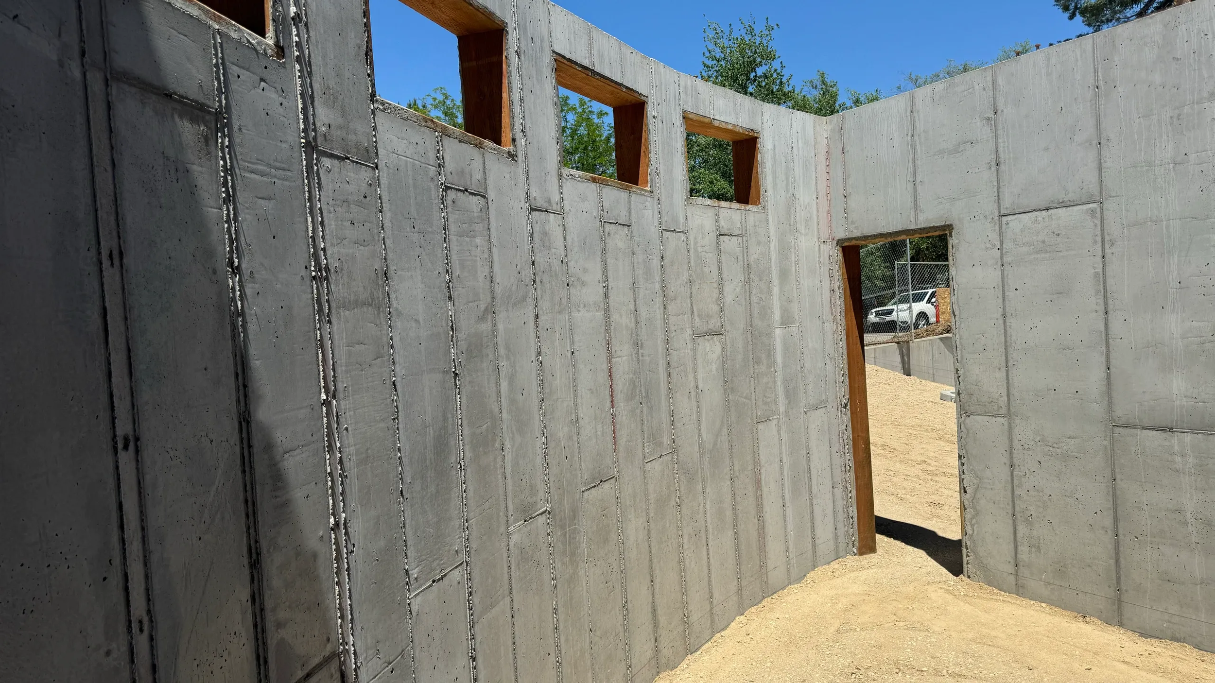 Concrete wall under construction with several window openings and a doorway, on a sandy ground with a chain-link fence and trees in the background.