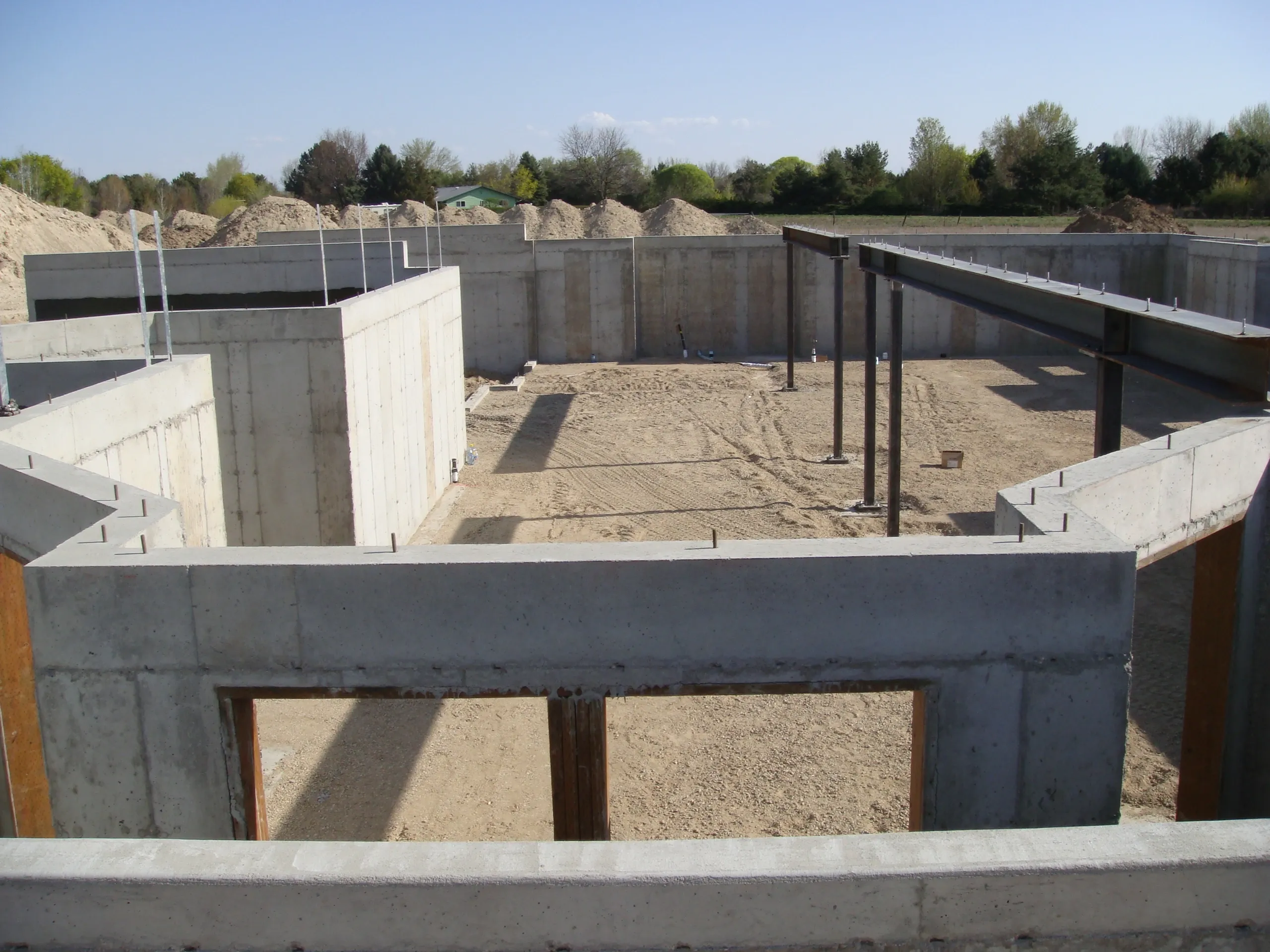 Construction site with concrete walls and metal beams, dirt ground, and scattered gravel, with trees and blue sky in the background.