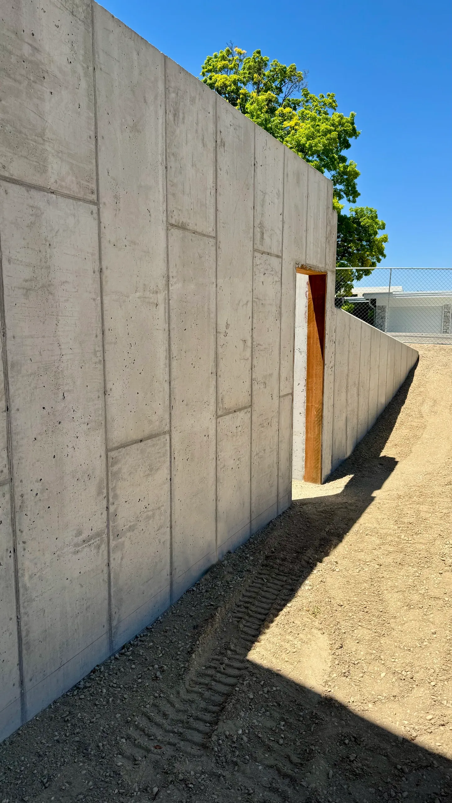 Concrete wall with an open doorway and a construction site outdoors, with a dirt ground and tire tracks, trees, blue sky, and a chain-link fence.