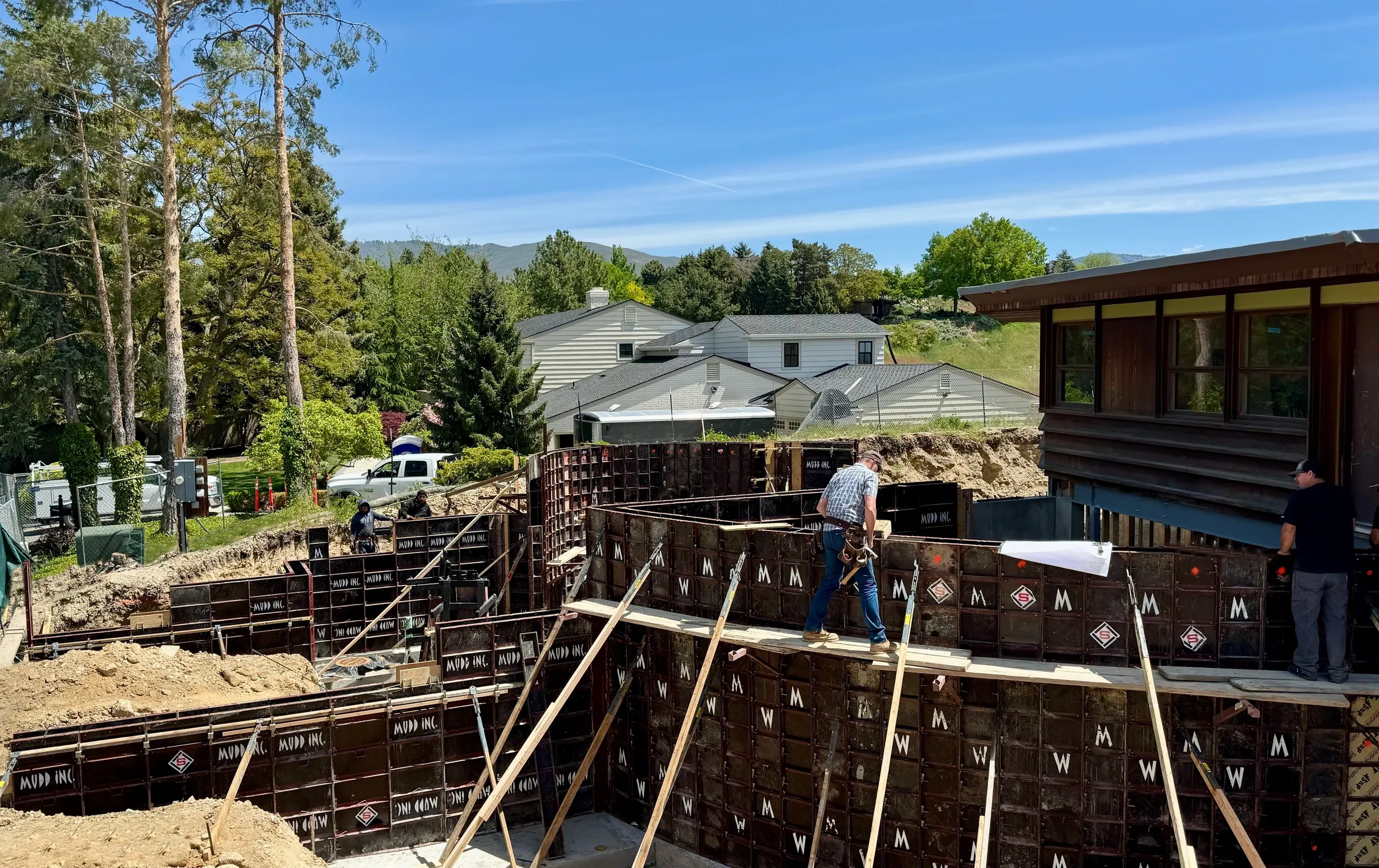 Construction workers building a foundation for a house with steel framing and wooden planks in a suburban neighborhood with trees and houses in the background on a sunny day.