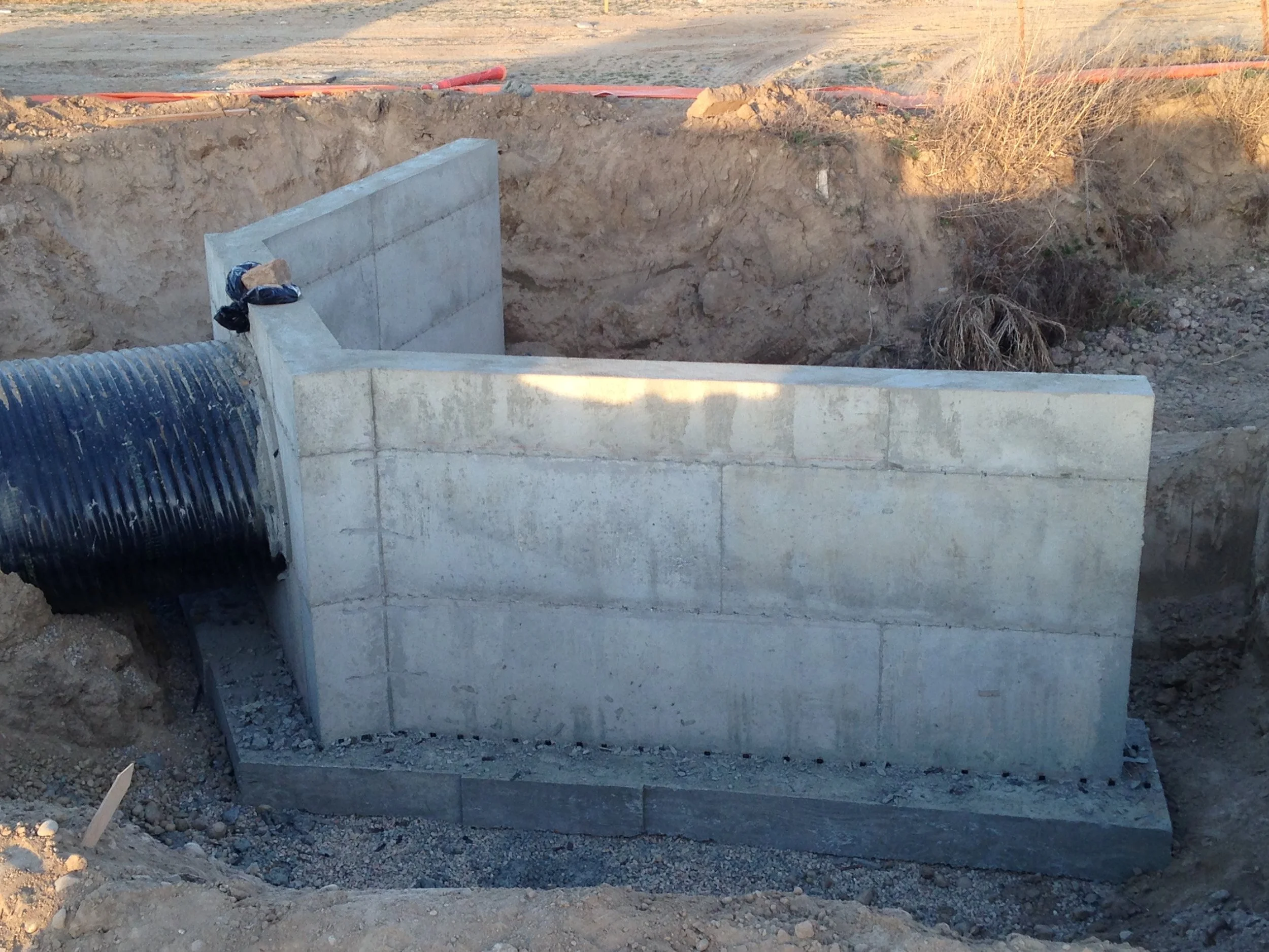Concrete foundation with a large black corrugated pipe protruding from the side, situated in a construction site with exposed dirt and surrounding dry vegetation.