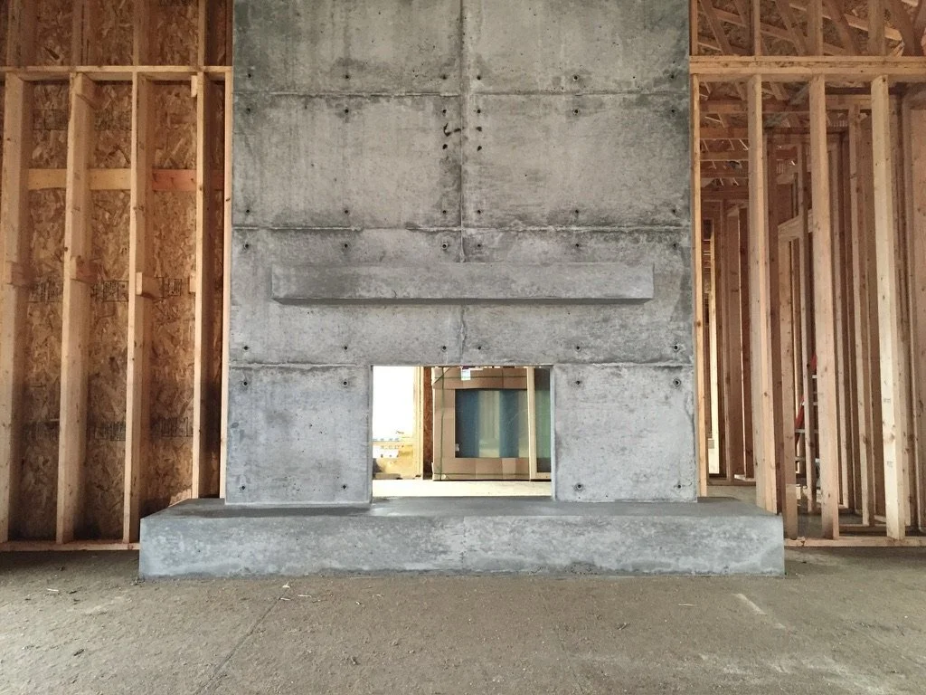 A concrete fireplace with a small opening, in a room under construction with exposed wooden framing and drywall.