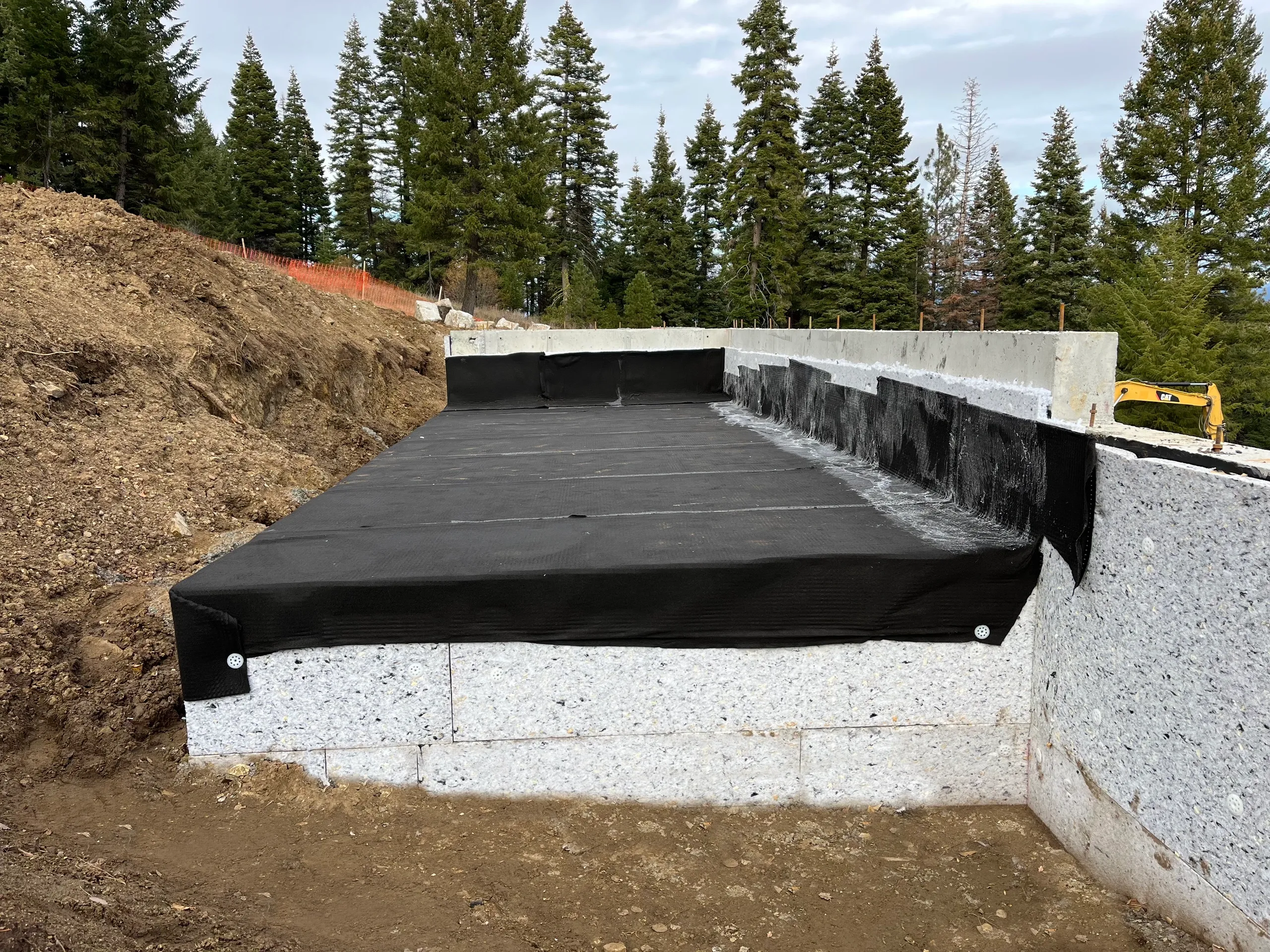 Construction site with a concrete wall and black waterproofing membrane, surrounded by dirt and trees in the background.