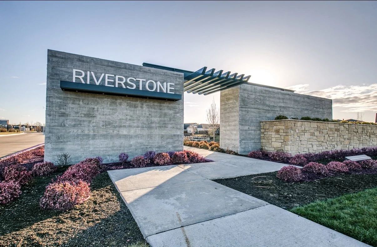 Modern entrance sign for Riverstone with concrete and stone walls, decorative plants and flowers, and a walkway leading into the community.