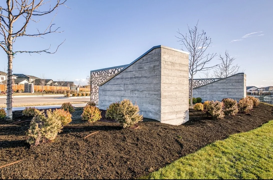Modern concrete wall with geometric design in a landscaped urban area with small bushes and leafless trees.