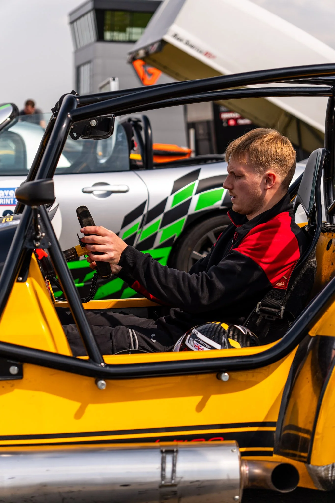 A young man with blond hair, wearing a red and black racing suit, is sitting in a yellow race car, holding the steering wheel. There is a helmet on the seat beside him and a car with checkered patterns visible behind him at a racing event with a buil