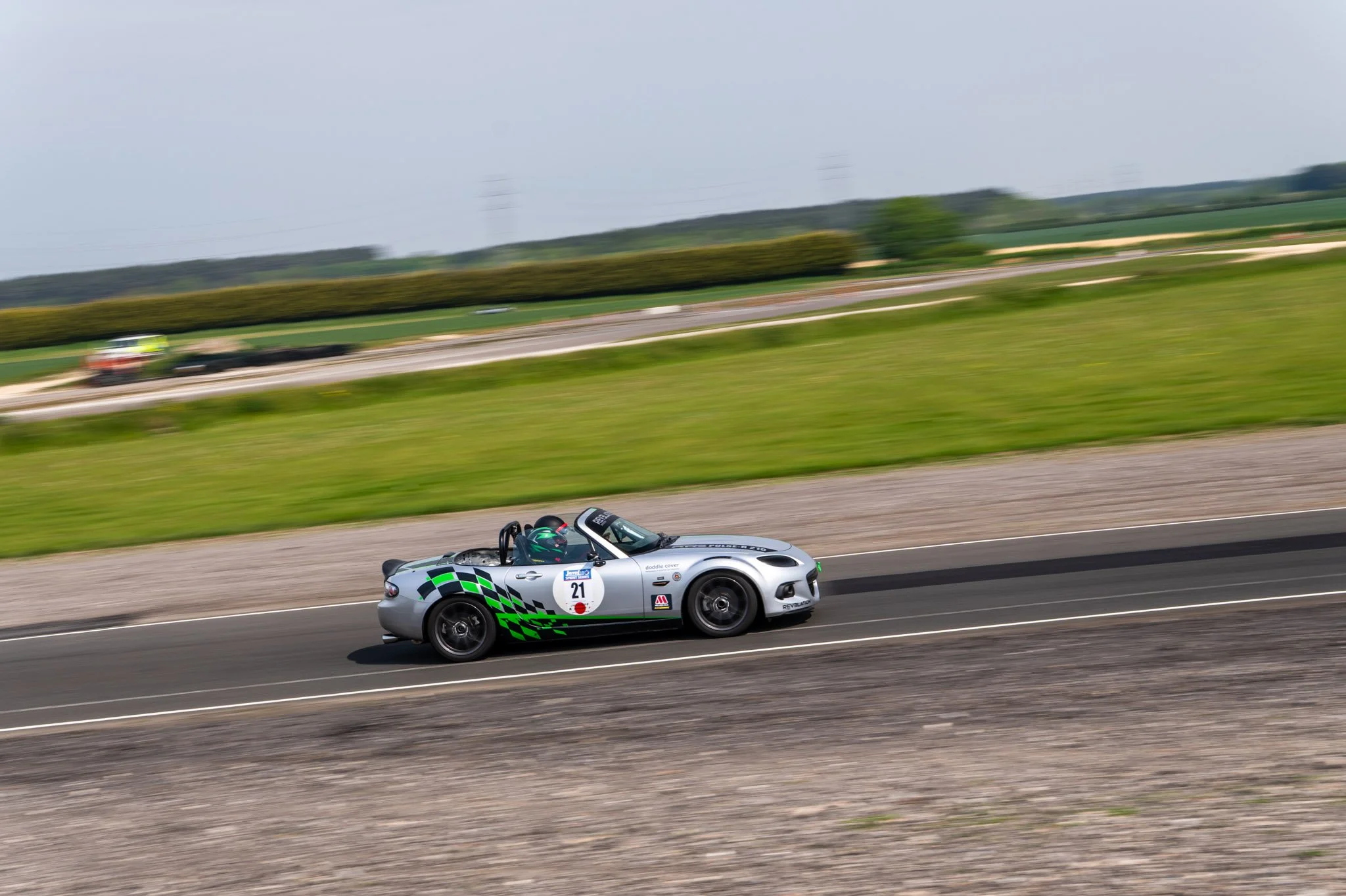 A silver racing car with green checkered pattern on the side driving on a race track, with blurred green grassy fields and patches of trees in the background.